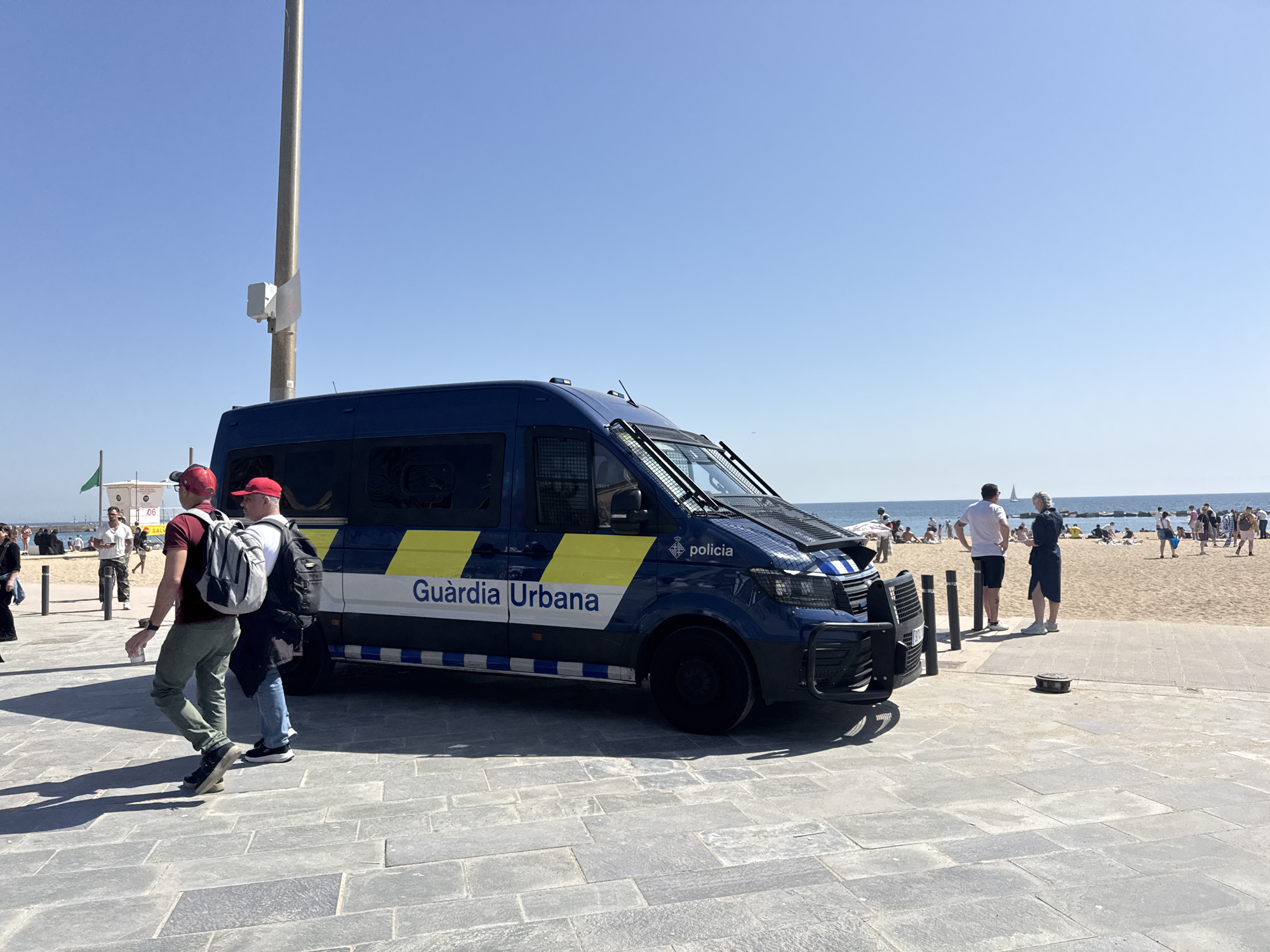 The image shows a blue "Guardia Urbana" (Barcelona's city police) van parked on a paved area adjacent to a beach in Barcelona, Spain. Two men, both wearing red baseball caps and backpacks, are walking past the van. The beach is crowded with people, and the sky is clear and blue. There are a few poles and flags visible in the background. The paved area appears to be part of a promenade or walkway along the beach.