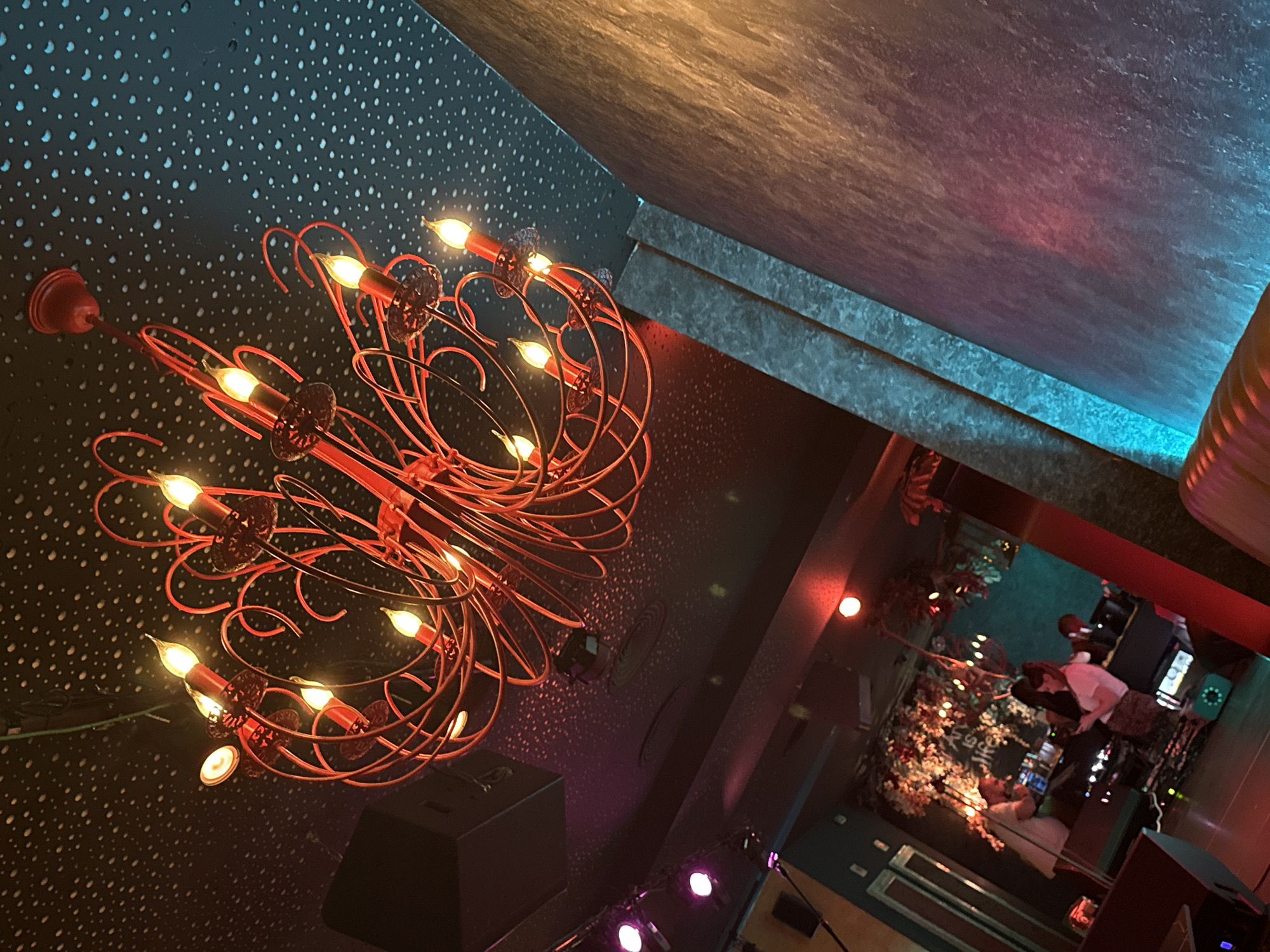 The image displays an interior scene from PM Club, a nightlife venue located in Sofia, Bulgaria, captured with an upward-tilted perspective. The dominant feature is a large, ornate red chandelier composed of intricate twisted and looping metalwork, suspended from the ceiling. It holds numerous candle-shaped light bulbs that emit a warm, amber glow.
The ceiling is dark, possibly black or dark grey, and uniformly covered with a pattern of small, irregular white speckles, resembling a starry night sky. In the upper left quadrant, a dark rectangular object, possibly a speaker, is mounted near the ceiling. Below it, purple and pink spotlights cast colored light onto the speckled ceiling.
On the right side of the image, a textured vertical wall, appearing to be made of stone or rough concrete, is prominently featured. A bright blue LED light strip illuminates a deep vertical crevice or edge in this wall, creating a stark blue line that contrasts with the warmer orange and pink light washing over the rest of the wall's surface.
In the lower background, an area resembling a bar or DJ booth is visible. A man with a beard, wearing a light-colored shirt, stands behind a counter, appearing to be operating some equipment. In front of the counter, a woman in a white top and a patterned skirt is engrossed in her mobile phone. The wall behind them is decorated with artificial foliage featuring white and red flowers, and a partially visible sign above reads "ART IS LIFE." The bottom edge of the image reveals a segment of an orange or tan padded bench or seating.