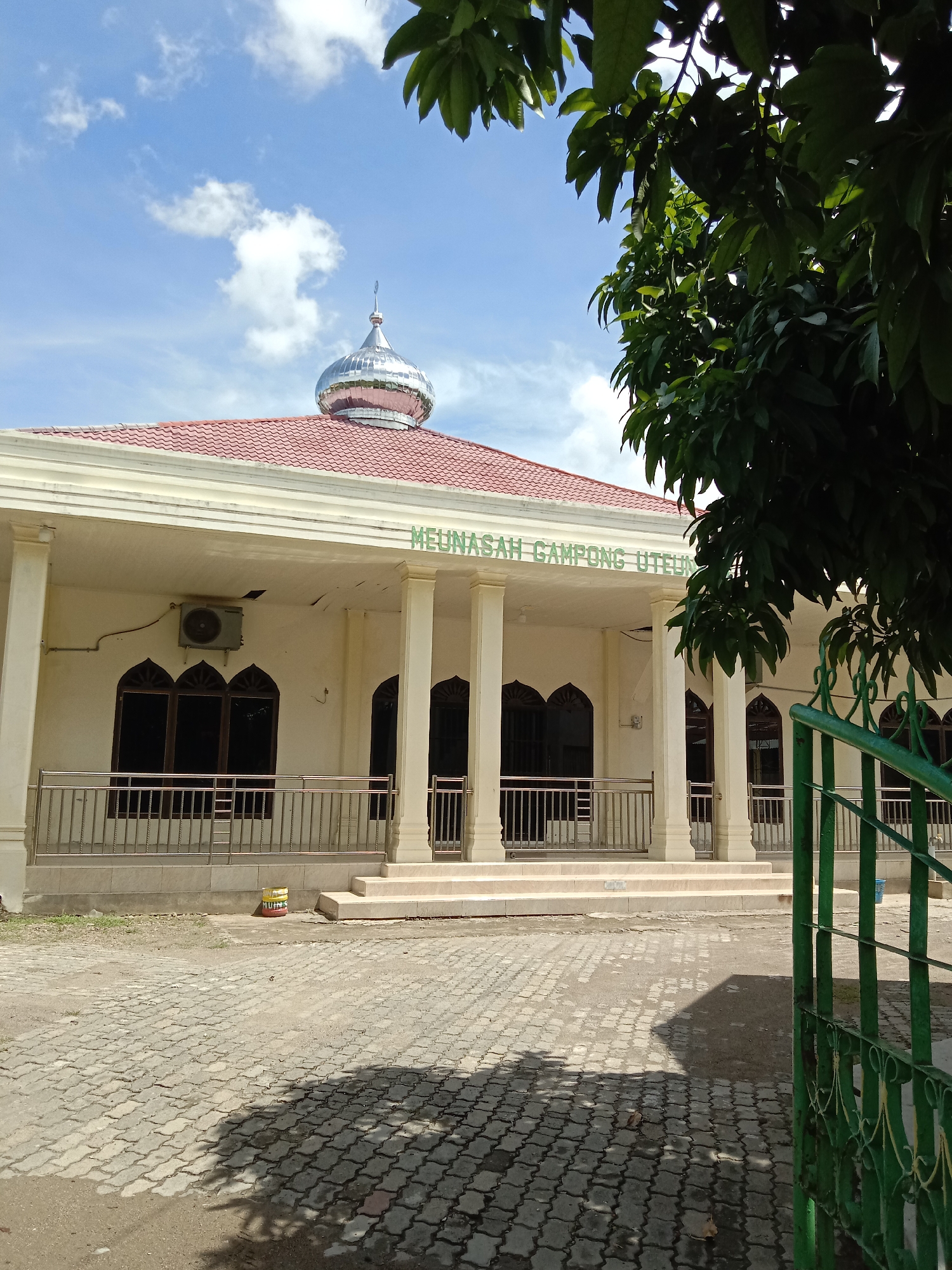 The image shows a mosque in Lhokseumawe, Indonesia, under a partly cloudy sky. The building is a light beige color with a red-tiled roof and a metallic silver dome. The facade features a series of arched windows with dark frames and a metal railing. The pillars are decorated with decorative bases. A green sign reading "MEUNASAH GAMPONG UTEUN" is present above the windows. An air conditioning unit is installed on the wall. The foreground features a paved area. A green metal fence is partially visible on the right side of the image. Trees are visible on the right side of the frame. A yellow can is sitting on the ground near the building. Shadows from the trees are cast on the paved area.