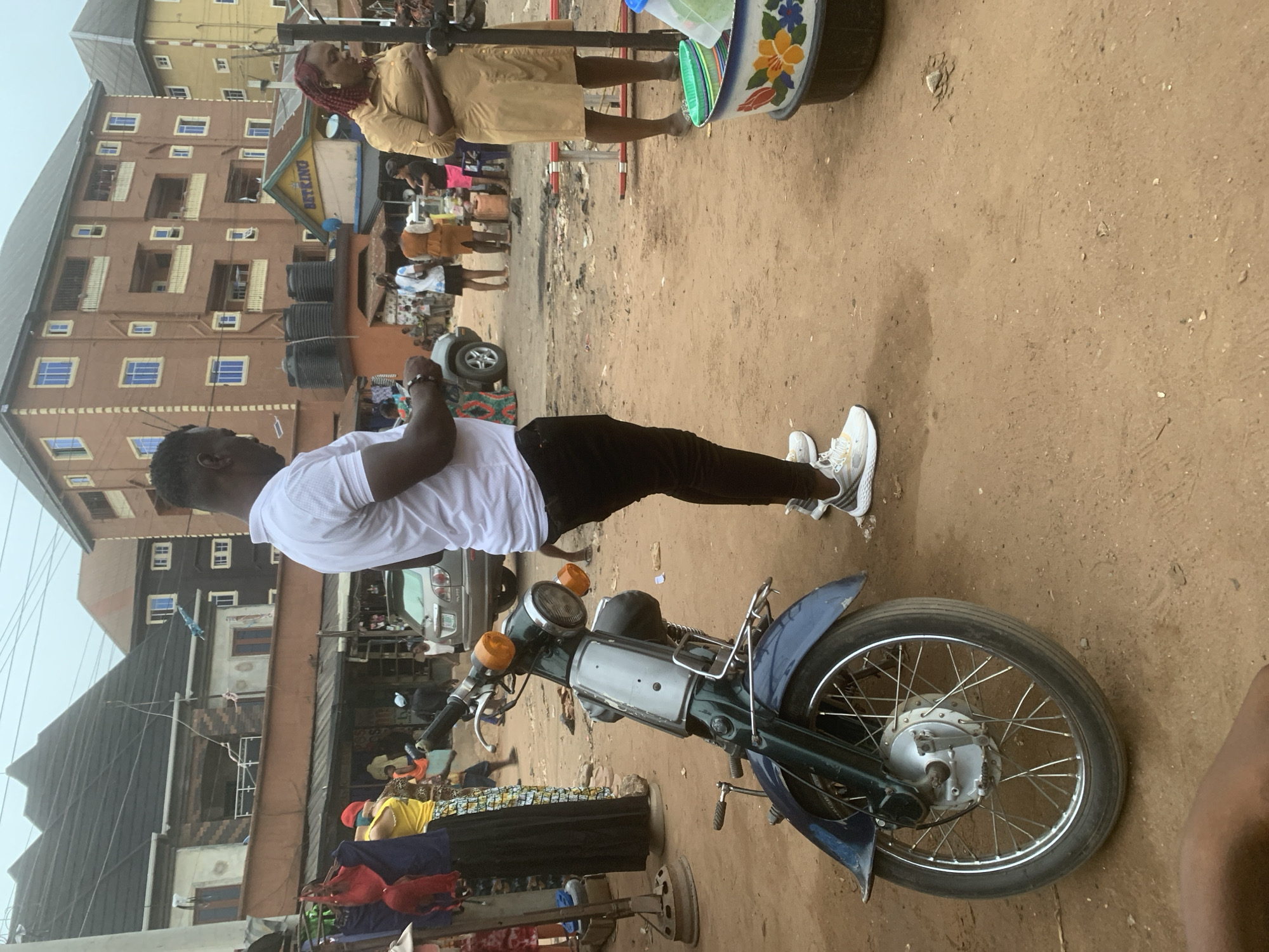 The image depicts a street scene in Onitsha, Nigeria. A man in a white t-shirt and black pants stands next to a motorcycle, facing a building marked "BEFKINO" with multiple windows and water tanks. Another person, wearing a brown outfit, is standing and selling items in the background. Other individuals and vehicles are visible in the distance. The ground is dirt.