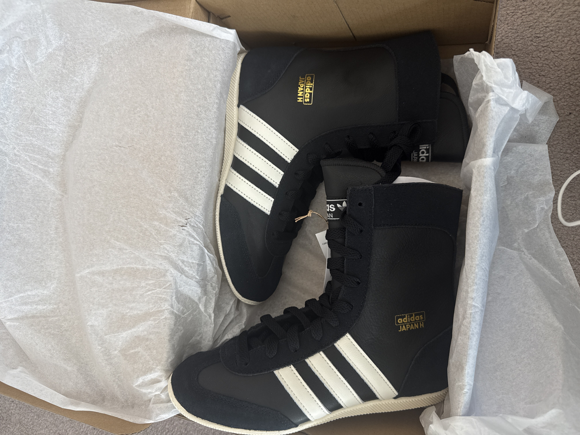 The image captures a top-down view of two new, black Adidas high-top shoes, resembling boxing boots, resting inside a brown cardboard shoebox. The shoes are predominantly black, featuring prominent white three-stripe branding on the sides and cream or off-white soles. Black laces secure the footwear.
Each shoe displays gold lettering on its side, clearly stating "adidas JAPAN H". A small, light-colored tag, likely a brand or product tag, is visible near the laces of the right shoe, showing the Adidas logo and partial text including "AN" and "ORIGINAL".
The shoebox is lined with white, crinkled tissue paper, indicating fresh packaging or an unboxing event. The surrounding background suggests an indoor setting, possibly a room with light-colored carpet. The lighting is bright and even, characteristic of daytime conditions. No individuals are present in the image. The scene conveys the fresh presentation of new athletic footwear, potentially in London, United Kingdom, as per the provided context.