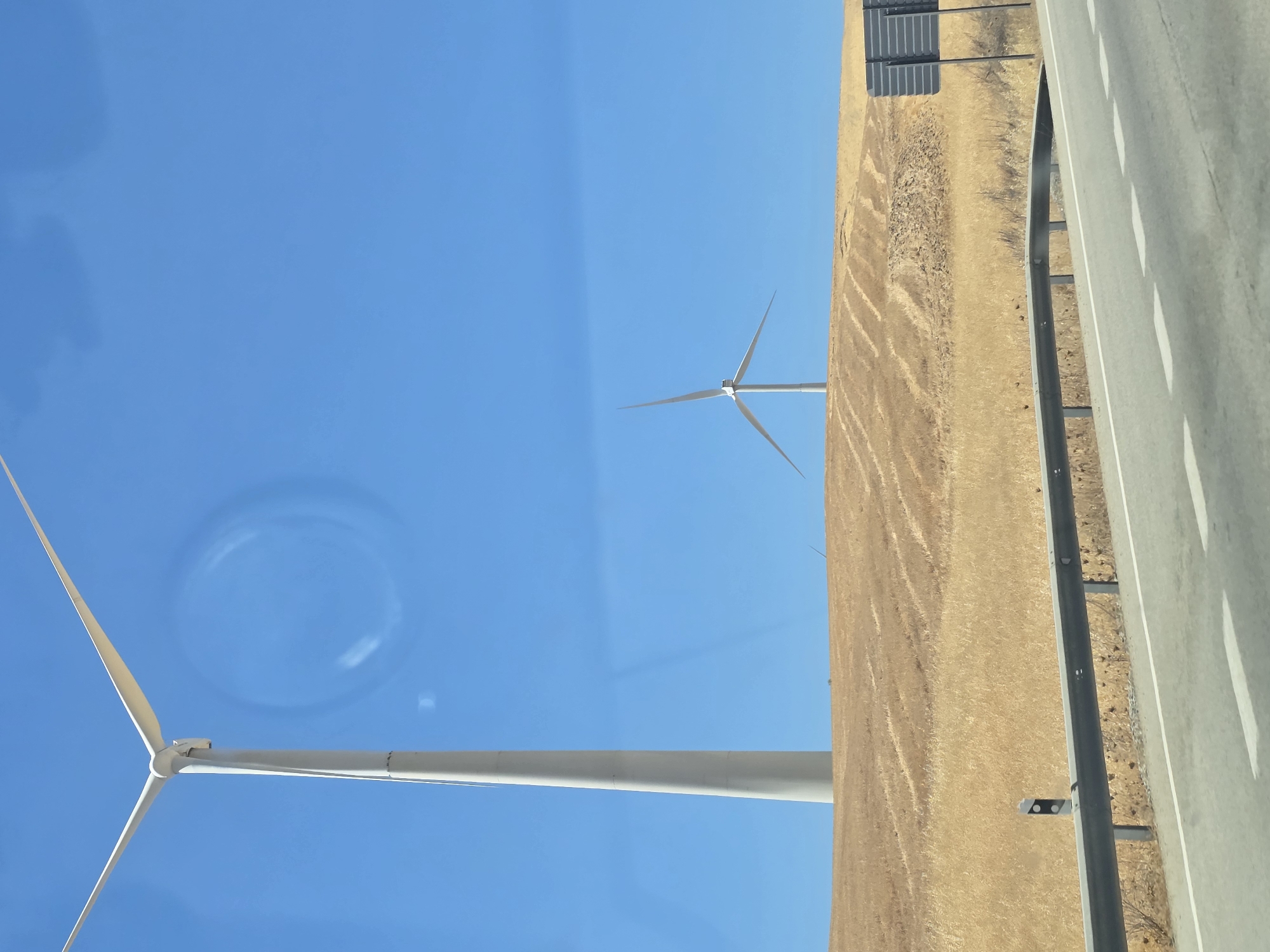 The image shows a roadside view with two wind turbines in the background, located in Teba, Spain. The foreground features a highway with white lane markings and a guardrail. Beyond the highway is a dry, golden field that appears to be harvested, with visible rows. The sky is clear and blue. The wind turbines are tall, white structures, one significantly larger and closer to the viewer than the other, which is situated further back on a slight rise in the terrain. The overall environment suggests a rural or semi-rural landscape.
