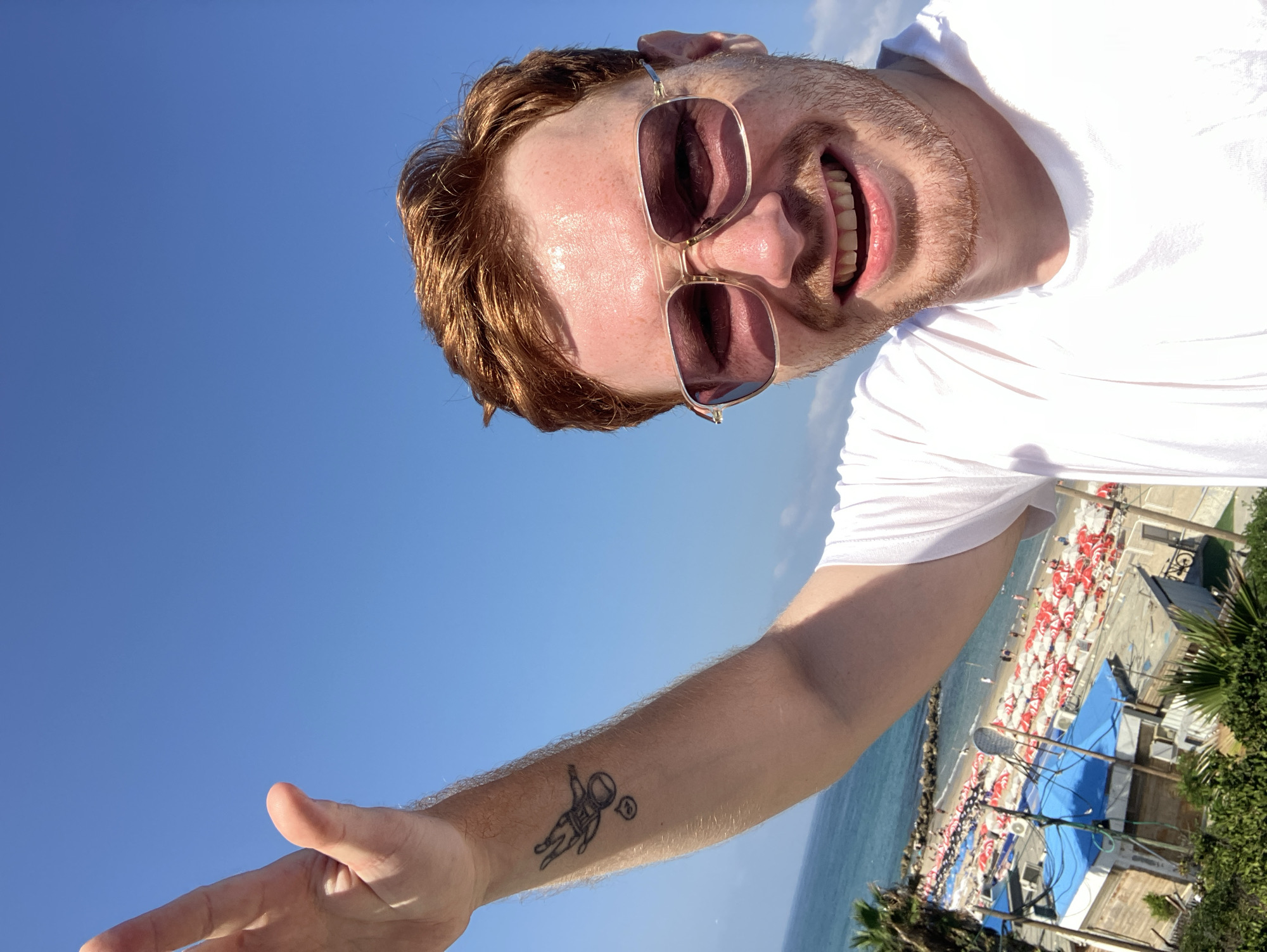 A man with red hair and stubble is taking a selfie at a beach in Bat Yam, Israel. He is wearing a white t-shirt and sunglasses. His left arm is raised, and he has a tattoo of an astronaut on his forearm. In the background, the sea, a beach with many umbrellas and people, and buildings can be seen under a clear blue sky.