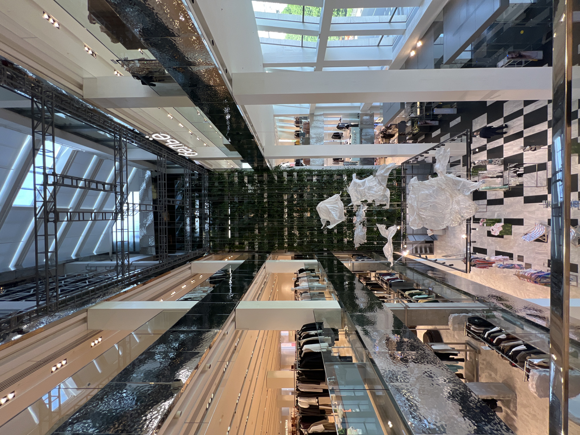 The image presents an unusual perspective, taken from directly above, looking down into the Central Department Store (TsUM) in Moscow, Russia. The building’s interior is modern and multi-level, with many floors visible. A system of mirrored surfaces reflects the interior elements, creating an Escher-like visual effect. The store sells clothing and accessories. There are hanging mannequins and other displays suspended from the ceiling. There are shoppers present within the store.
