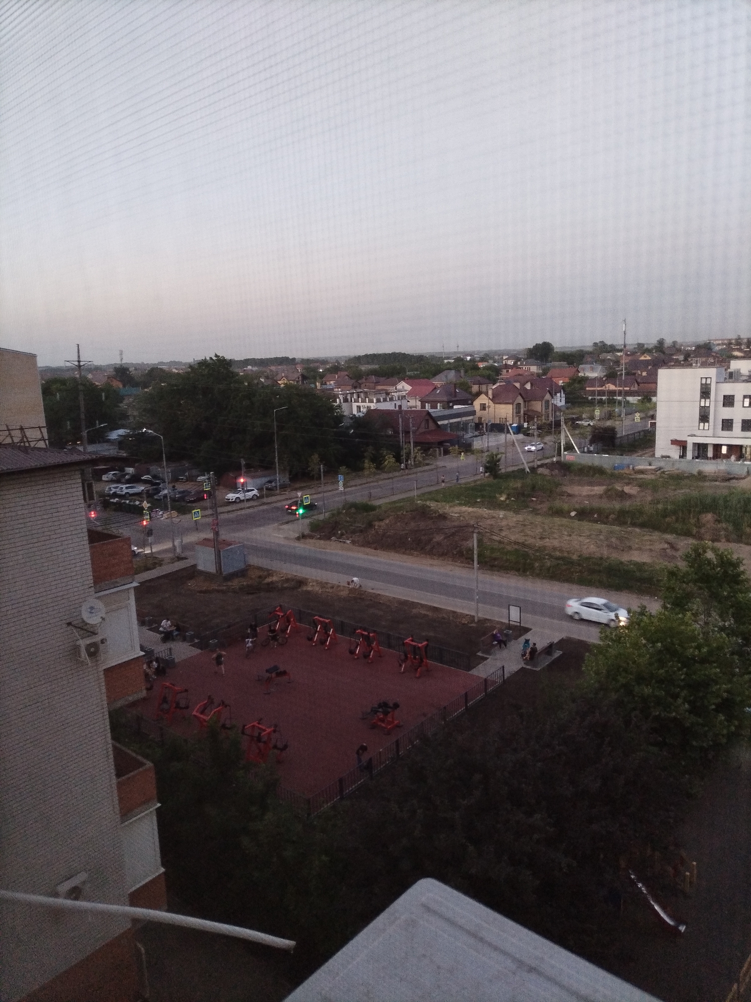 This is an aerial, eye-level view of a residential area in Rossiyskiy, Russia, captured from a building. The scene shows a street intersection with traffic lights, several cars, and a playground with red exercise equipment. A multi-story building is visible on the left, and a row of houses with red roofs stretches across the horizon. The sky is overcast, and the overall lighting suggests it is either dawn or dusk. There are a few people visible, some on the playground and some walking on the sidewalk.
