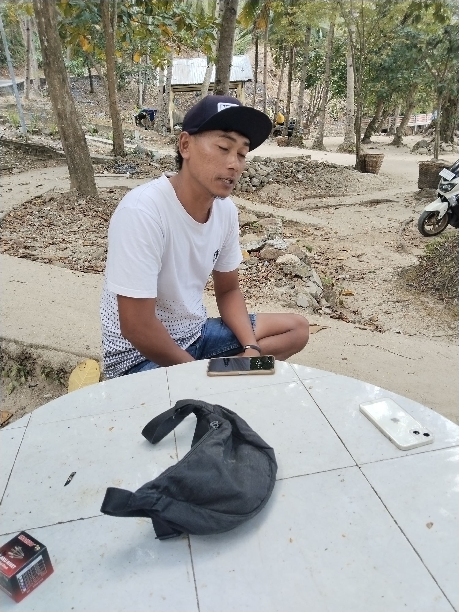 A man wearing a white t-shirt, denim shorts, and a black baseball cap sits at a white tiled table outdoors. He appears to be speaking or singing, with his mouth open and eyes closed. On the table are a black fanny pack, a smartphone, and a box of rivets. In the background, a white motorcycle is parked, and there are trees and a rustic building with a metal roof. The ground is sandy and covered with leaves and debris. The scene suggests a casual, outdoor setting in a rural or semi-rural environment, possibly near a beach or a village. The presence of the building and motorcycle indicates a residential or commercial area. The overall atmosphere is relaxed and informal, with the man seemingly engaged in a personal moment.
