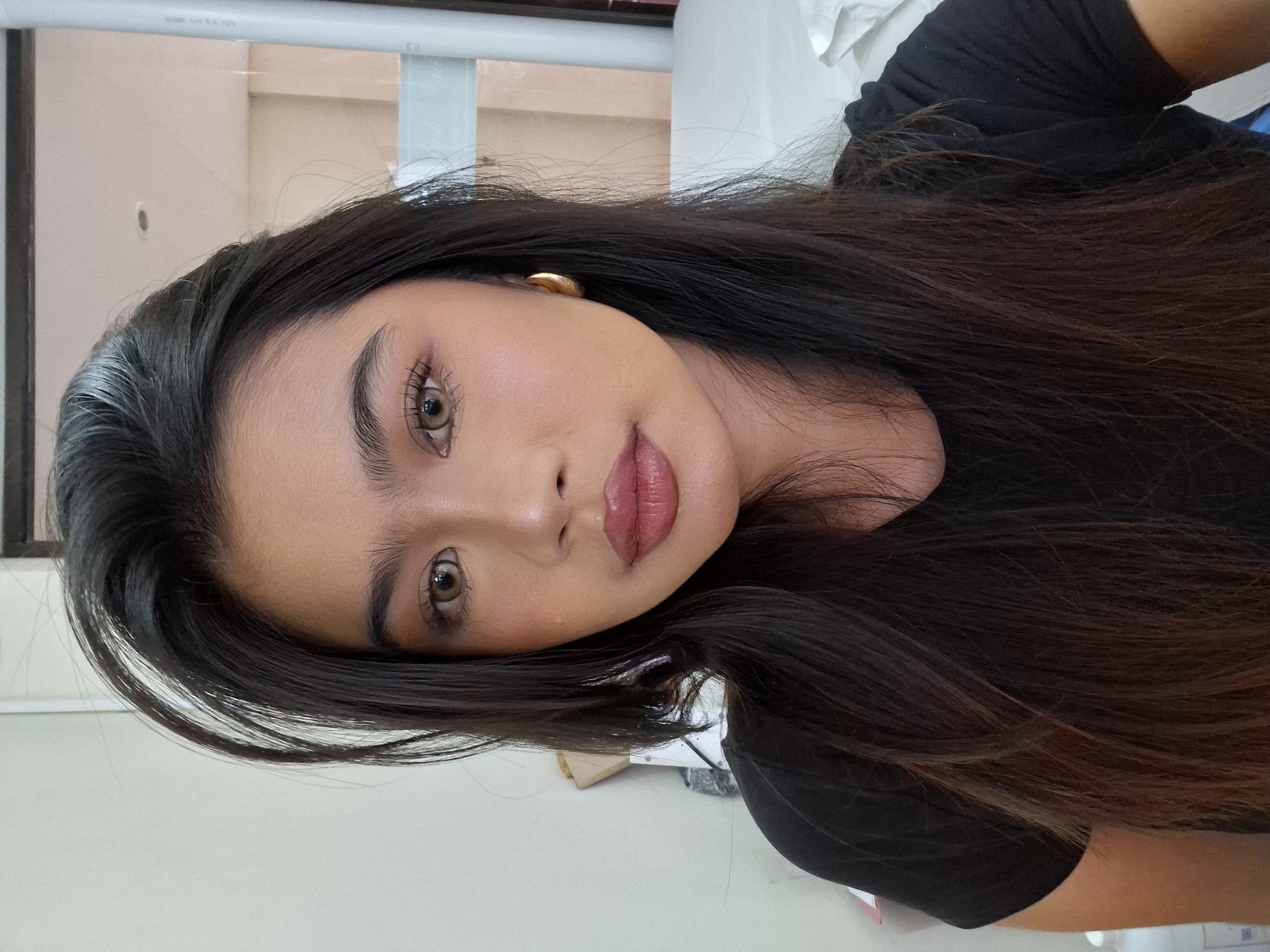A young woman with dark hair and makeup poses for a selfie in a room. She is wearing a black t-shirt and a gold hoop earring in her right ear. The background appears to be a bedroom with a white bed and some boxes on a shelf. The lighting is natural, coming from the left side of the frame. The image quality is good, with a shallow depth of field that blurs the background.