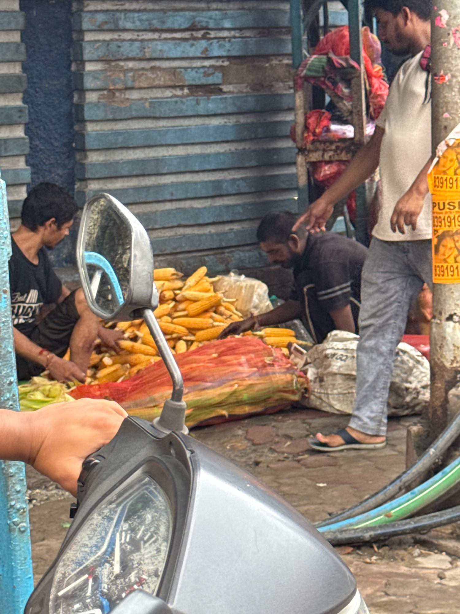 The image depicts a street scene, likely in Kolkata, India, with a focus on a market stall or roadside vendor. In the foreground, the handlebars and dashboard of a grey scooter are visible, with a person's hand resting on the handlebars. To the left, a blue painted column is present. Behind the scooter, a pile of yellow corn cobs is displayed on the ground, partially covered by colorful fabrics, suggesting a produce market.
Several individuals are present. On the left, a person wearing a black t-shirt and shorts is partially obscured, tending to the corn. In the center, another person wearing a dark shirt is crouched down, also interacting with the corn. To the right, a person in a light-colored t-shirt and grey jeans stands, pointing towards something not entirely in frame, possibly indicating a sale or interaction with a customer. A rustic shelving unit with red and green fabrics is visible behind the standing individual. The background consists of a textured, weathered wall with horizontal blue and grey stripes, typical of older urban structures in Kolkata. The ground is paved with brick or stone. The overall atmosphere suggests a busy, everyday market environment.