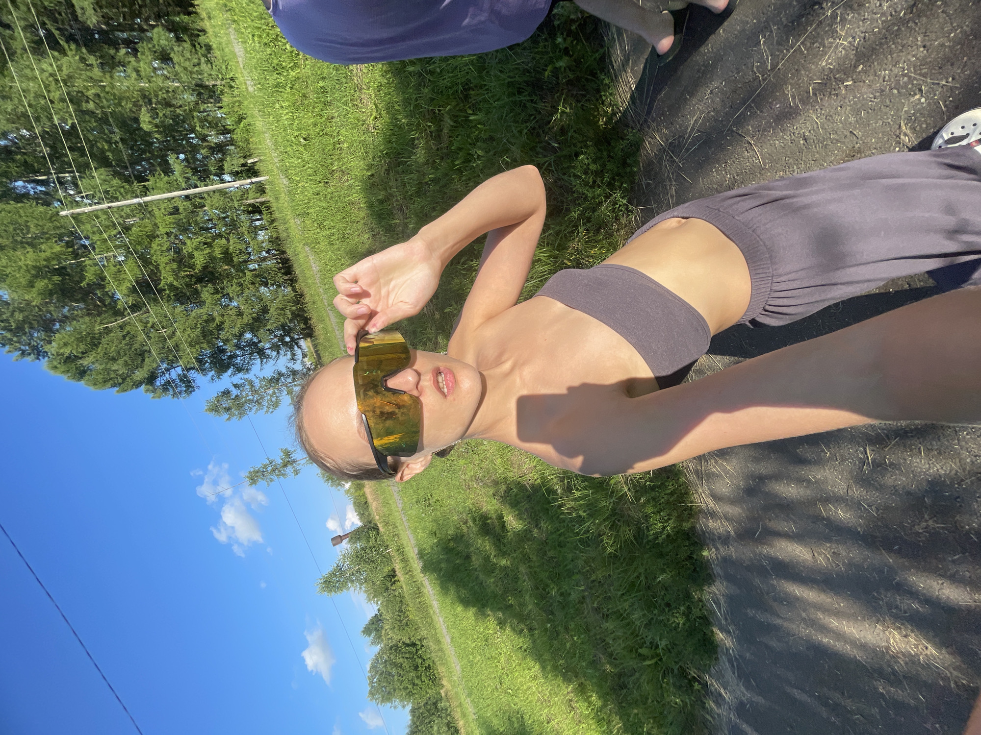 This media file features a young person, likely female, wearing large, reflective sunglasses and a dark, strappy top or sports bra. They are also wearing loose, dark-colored athletic pants. The person is positioned outdoors in what appears to be a rural or semi-rural setting.
The scene is characterized by a bright, sunny day with a clear blue sky dotted with a few white clouds. The ground is a mix of dark, dry dirt or gravel and lush green grass, suggesting a warm, possibly summer, day. In the background, there are tall trees and what appear to be power lines supported by wooden poles. A wooden structure, possibly a fence post or part of an old building, is also visible in the distance.
The person is posing for the camera, holding the edge of their sunglasses with one hand, and looking towards the lens with an open-mouthed expression. The lighting suggests it is likely midday or early afternoon, given the bright sun and the sharp shadows cast on the ground. The overall mood appears casual and possibly energetic, fitting for an outdoor activity. There is no visible text in the image. The location context provided indicates this scene is in Podshivalovo, Russia.