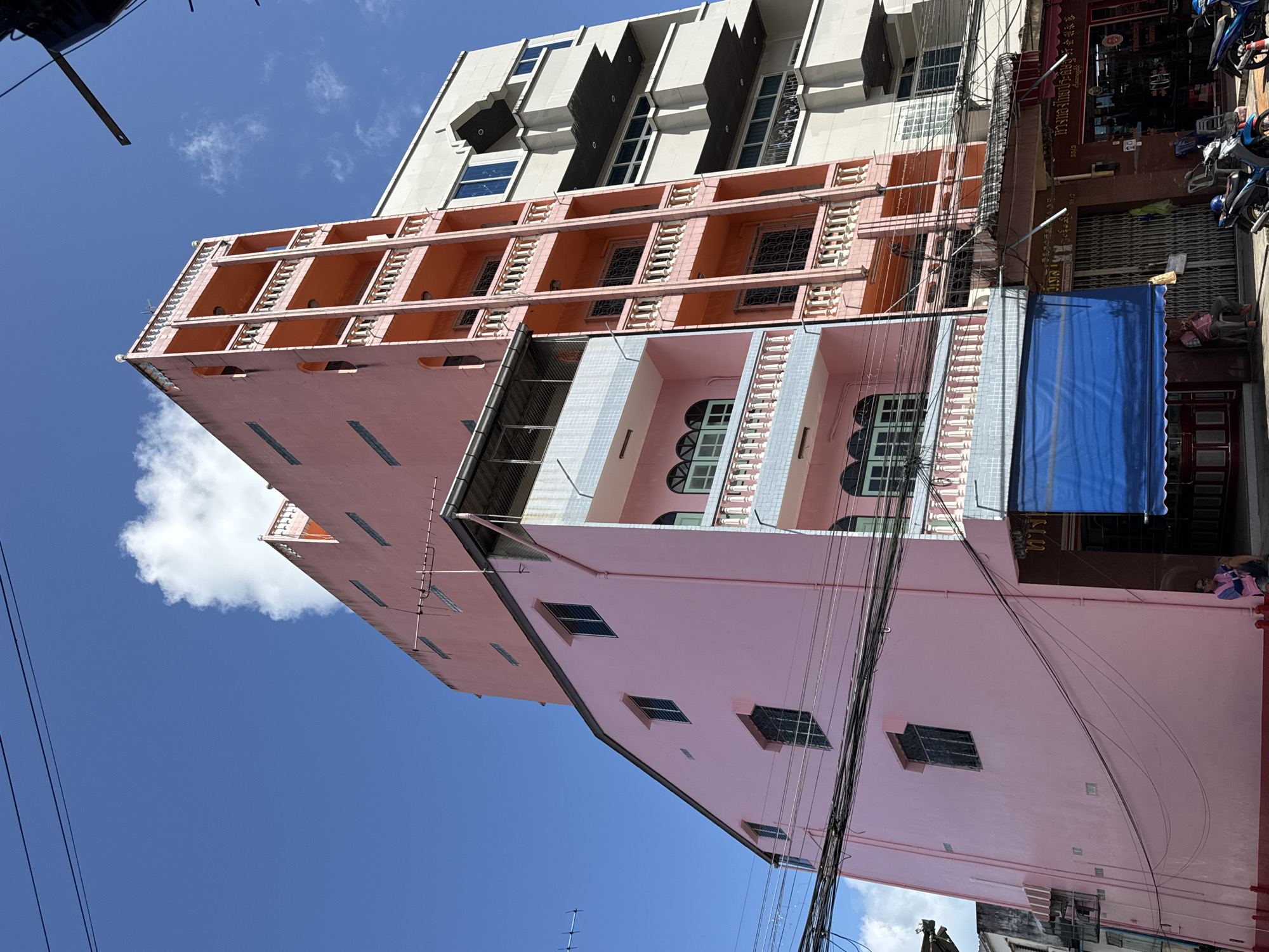 This image depicts a street scene in Ranong, Thailand, featuring a row of multi-story buildings. The primary building on the left is a narrow, tall structure painted a bright pink, with a sloped roofline and several windows. Adjacent to it is a taller, salmon-pink building with a prominent vertical section featuring multiple balconies with white balustrades and arched window openings. To the right of this is a lighter-colored building, possibly cream or beige, with a more modern facade and a series of horizontal dark-colored overhangs or balconies. The buildings are situated along a street with overhead power lines crisscrossing the frame. A blue awning is visible on the ground floor of the pink building. In the foreground, several motorcycles are parked. Two individuals are partially visible: one sitting on a ledge in front of a storefront on the ground floor of the pink building, and another person near the motorcycles. The sky is a clear, vibrant blue with a few scattered white clouds. The architecture suggests a densely populated urban environment, likely a commercial or mixed-use area.