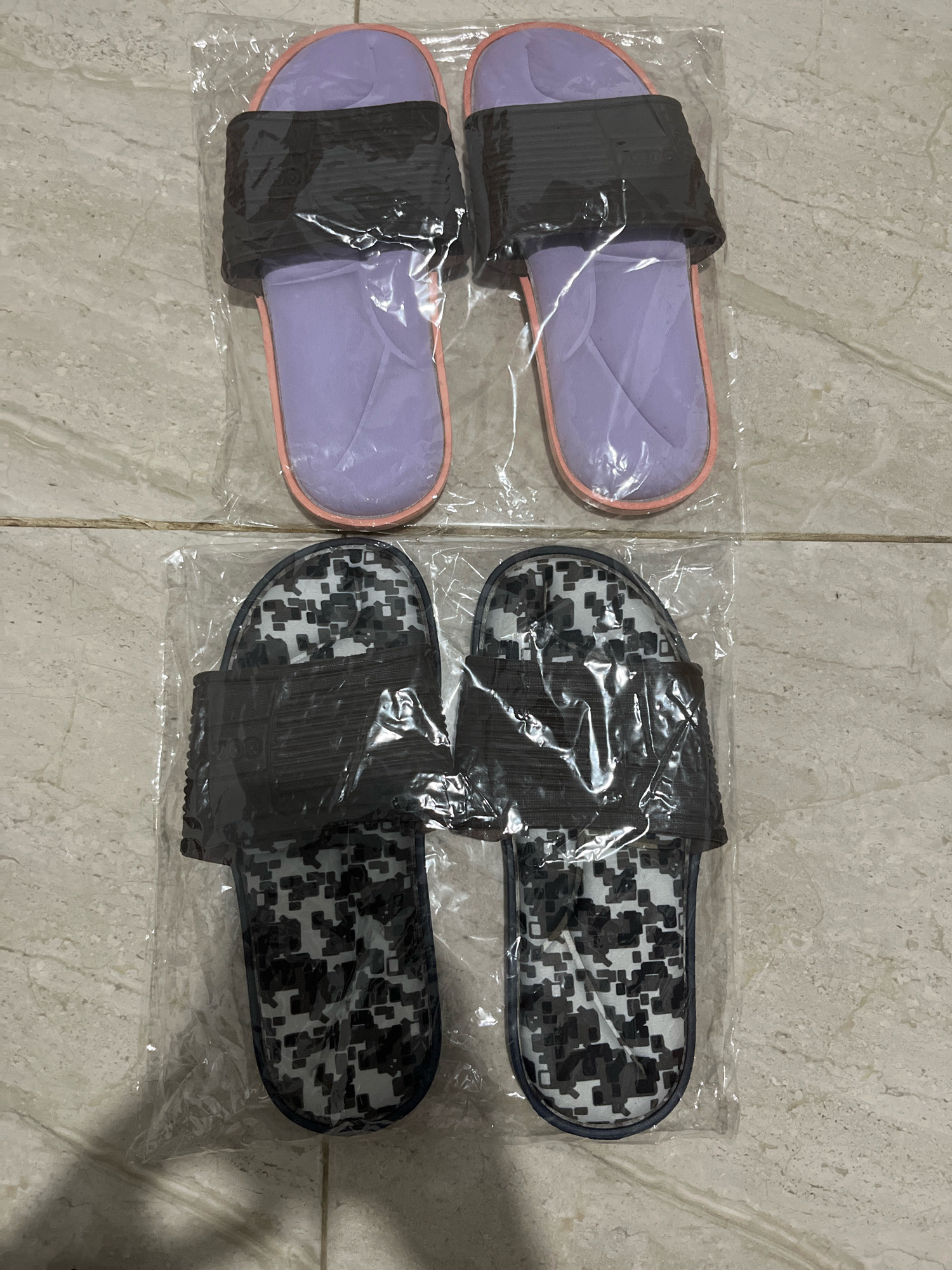 The image shows two pairs of slippers on a tiled floor. The top pair has lavender insoles and dark grey straps. The bottom pair has a digital camouflage pattern on the insoles and dark grey straps. Both pairs are individually wrapped in clear plastic. The slippers are arranged in two rows, with each pair occupying a horizontal space. The background is a neutral-colored tiled surface with visible grout lines. The lighting is overhead, casting slight shadows. There are no people or any other objects present in the scene.