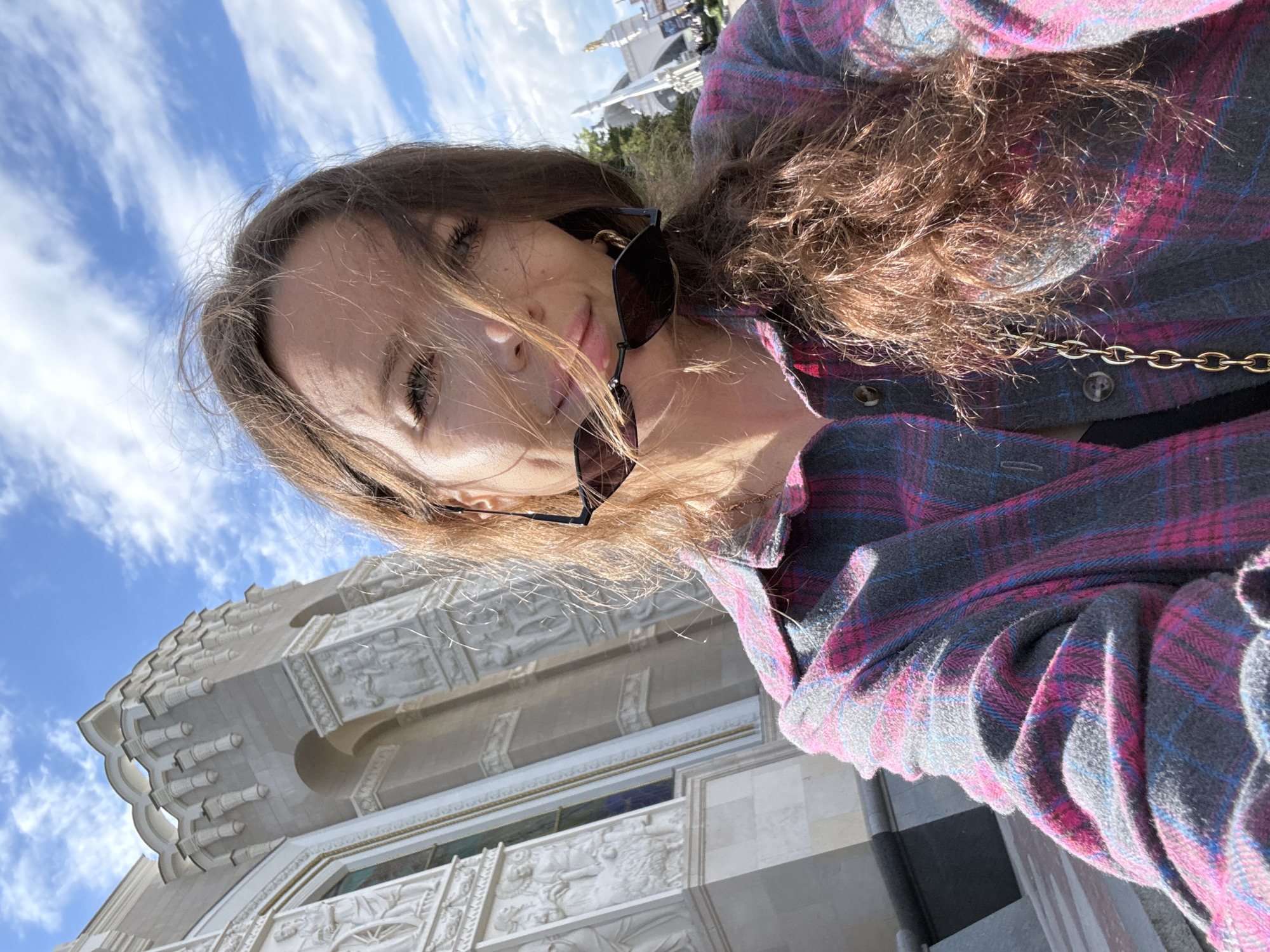 A young woman with long, brown, wavy hair is taking a selfie outdoors. She is wearing sunglasses on her nose and a pink and blue plaid shirt with a gold chain accessory. Her hair is partially obscuring her face, and she has a slight smile.
The background shows a bright blue sky with scattered white clouds. To her left and below, a detailed white stone building with intricate carvings and architectural elements is visible. The building's design suggests it could be a historical or decorative structure. In the upper right corner, a glimpse of another white structure, possibly a monument or tower, can be seen against the sky.
The lighting and clear sky indicate it is daytime, likely sunny. The overall mood appears cheerful and relaxed, typical of a tourist or someone enjoying an outing. The context provided places the scene in Moscow, Russia. There is no visible text in the image.