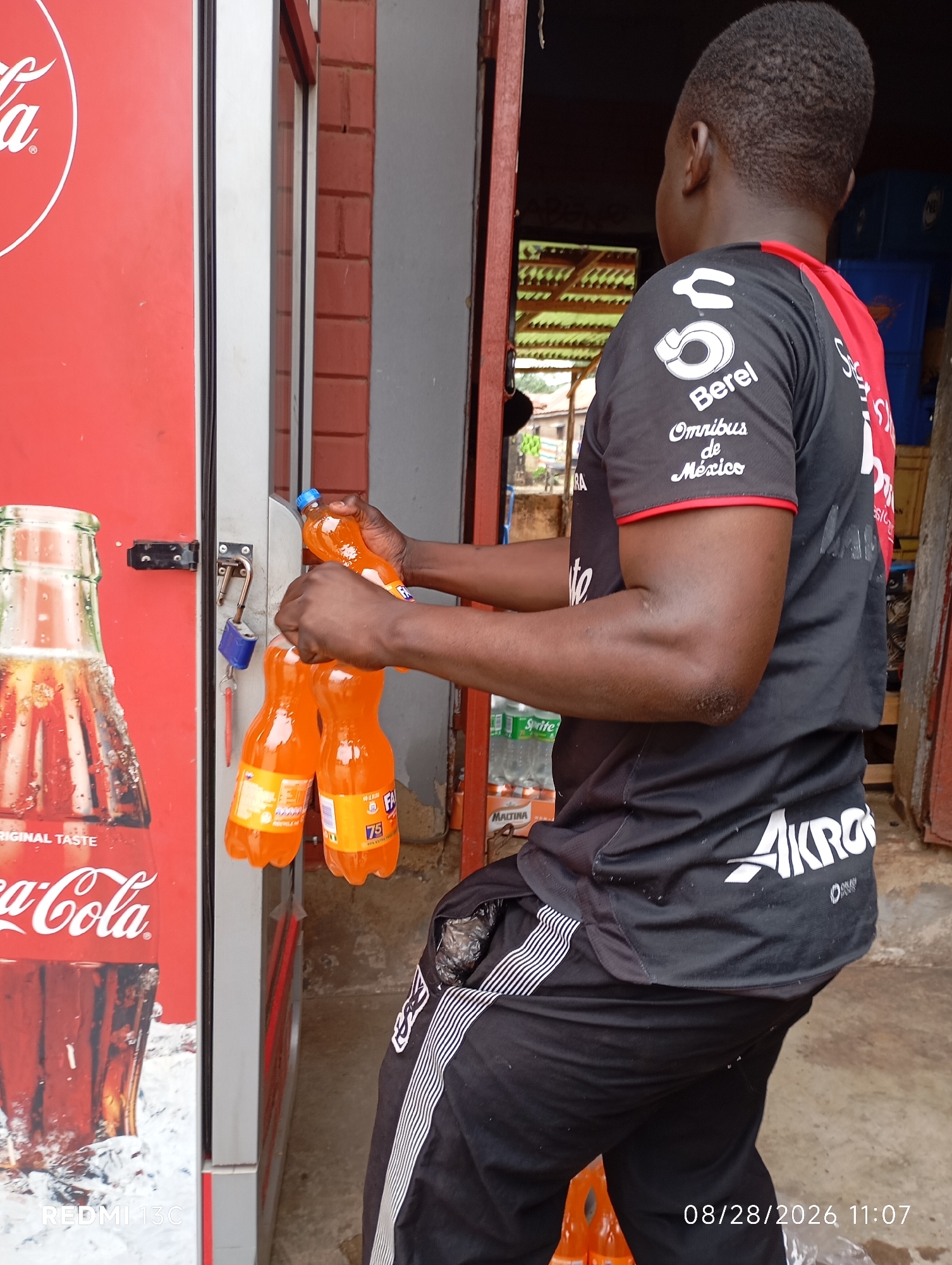 In the city of Shendam, Nigeria, a young man wearing a black jersey with red accents and black track pants is interacting with a vending machine. The man is holding two orange-colored bottles of soda, possibly Fanta, which appear to be just dispensed from the machine. The vending machine itself is predominantly red with a Coca-Cola advertisement visible on its left side. A lock is attached to the machine's access point, suggesting it is secured. The background reveals a brick wall and other elements of an outdoor market or shop setting. The timestamp "08/28/2026 11:07" is overlaid on the image.