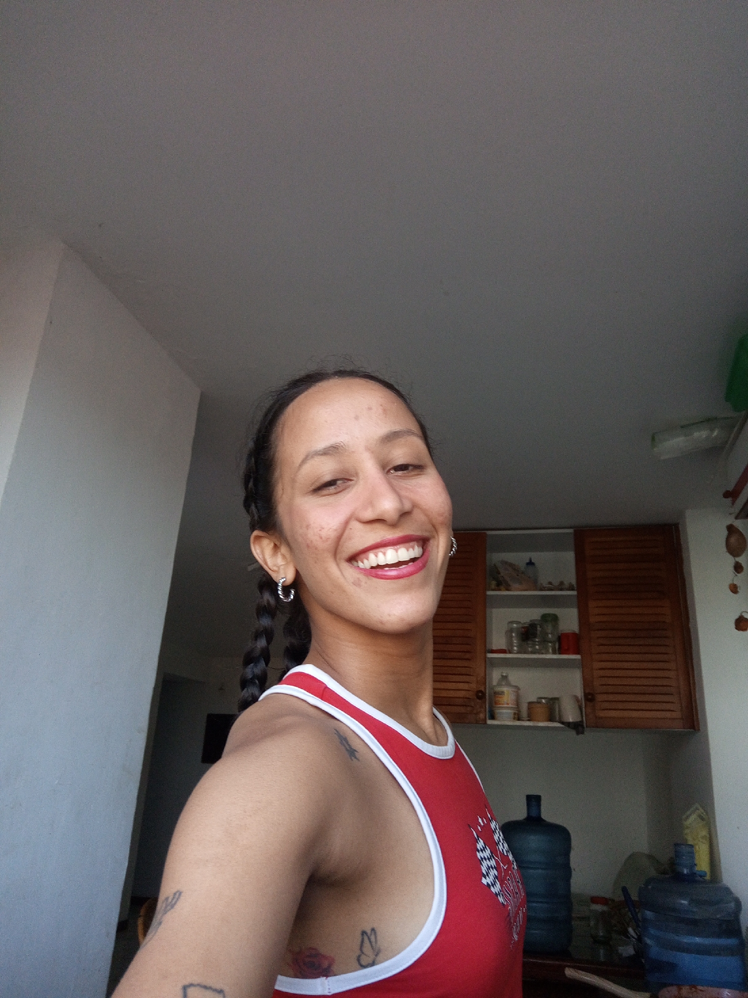 A woman with dark braided hair, wearing a red tank top with white trim and a checkered flag graphic, smiles at the camera. She has hoop earrings in her ears and visible tattoos on her arm and torso. She is positioned in what appears to be a kitchen. In the background, there are wooden cabinets, shelves stocked with jars and containers, and large blue water jugs. The overall scene suggests an indoor domestic setting, possibly in Porlamar, Venezuela, given the presence of large water jugs which are common in areas with potential water supply issues. The lighting suggests it is daytime.