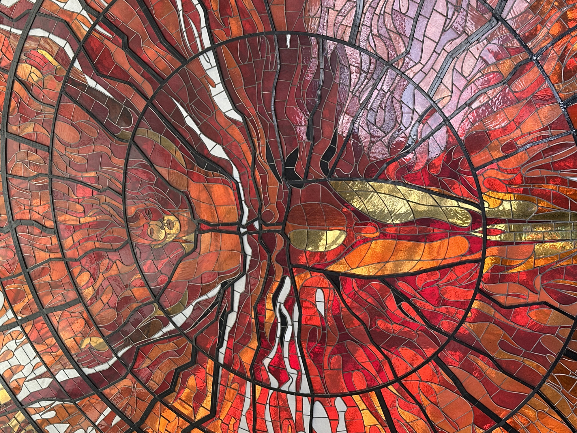 This is a detailed close-up photograph of a stained-glass mosaic dome. The design features a circular framework divided into sections by dark lines. The glass pieces within these sections are predominantly in shades of red, orange, and gold, creating a warm, vibrant effect. The pattern within the dome appears abstract, with some sections suggesting organic shapes or flames. Some elements include areas of white glass. This mosaic dome is located in Toluca, Mexico.