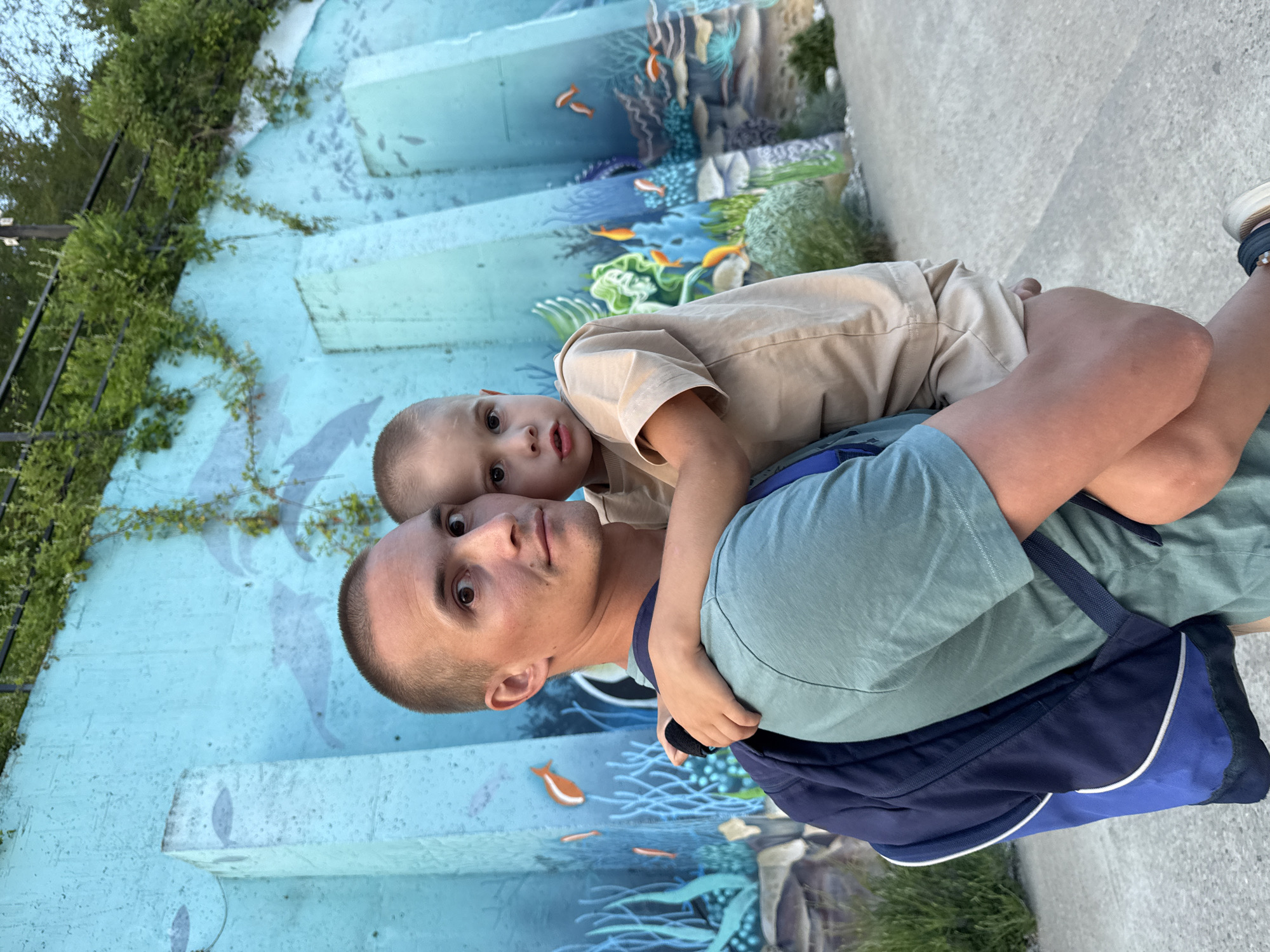 A man in a teal t-shirt and a blue backpack carries a young boy in a tan t-shirt in front of a blue mural of an underwater scene. The mural depicts fish, coral, and seaweed. The man has a short buzz cut and is looking towards the camera with a surprised expression. The boy is looking at the camera with a neutral expression. The mural is on a concrete wall, and there is a fence with greenery overhead. The ground is a paved surface. The image appears to have been taken outdoors during daylight. It is plausible that this is a public space in Nebug, Russia, given the context.
