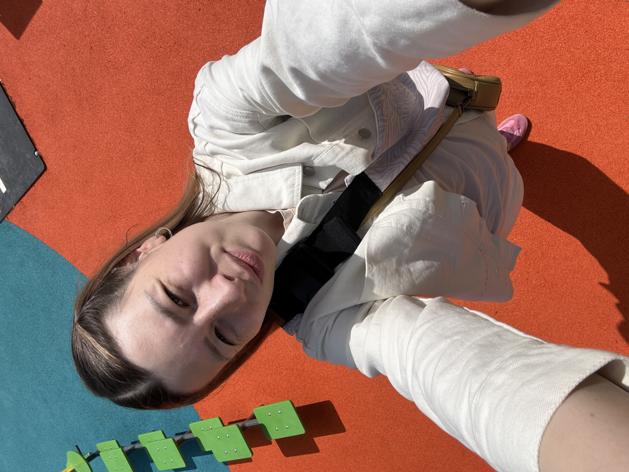 A woman is taking a selfie outdoors. She is wearing a white jacket and a small khaki-colored purse. She is on an orange and blue playground with green plastic stepping stones. The location is potentially a playground in Moscow, Russia.