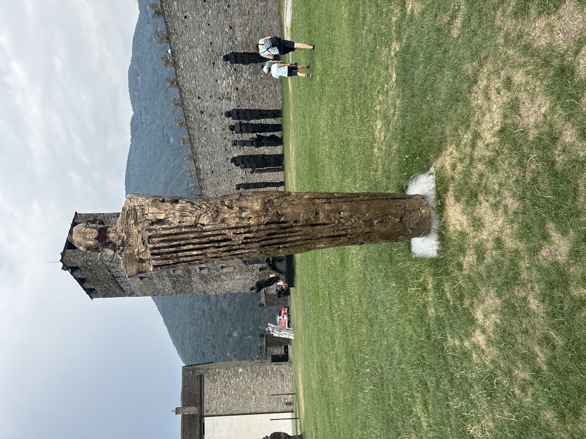 The image depicts an outdoor scene at Castel Grande in Bellinzona, Switzerland. In the foreground, a tall, bronze-colored abstract sculpture stands on a grey base in a grassy area. Behind the sculpture, a stone castle wall with battlements stretches across the middle ground. To the left of the sculpture, part of a stone building with a white facade is visible. Further back, to the right of the sculpture and in front of the castle wall, are several dark, slender figures, resembling statues or sculptures. Two individuals, a woman and a child, are walking away from the camera towards the castle wall on the right side of the image. The child is wearing a blue shirt and a white hat, and the woman has a backpack. In the distance, mountains are visible under a cloudy sky. A red and white car is parked in the background near an entrance to the castle grounds. The overall environment is that of a historical site and sculpture park.