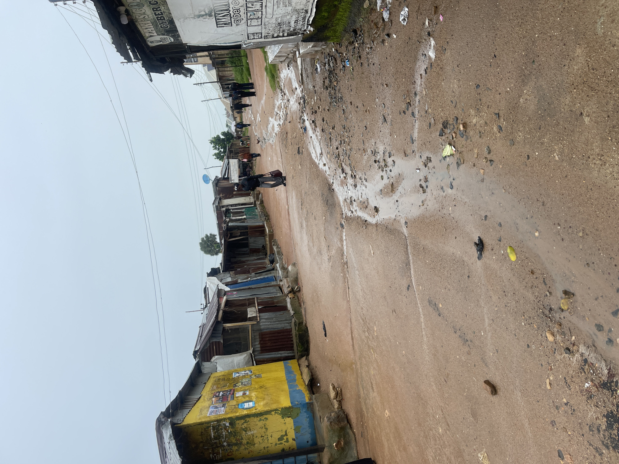 This is a street scene in Rafin Par, Nigeria. The street is unpaved and muddy, with puddles and debris scattered across its surface. On the left side of the street are several small shops or stalls constructed from corrugated metal and wood, some adorned with posters. One prominent stall is painted yellow and blue. On the right side of the street, more shops are visible, one with a sign that reads "ZAMISCO CYBER C". Several people are walking along the street. One person in the foreground is carrying a red bag and wearing dark clothing. Further down the street, a small group of people are walking away from the camera. A chicken is visible in the middle of the road. The sky is overcast, suggesting a cloudy or rainy day. Power lines are strung overhead.