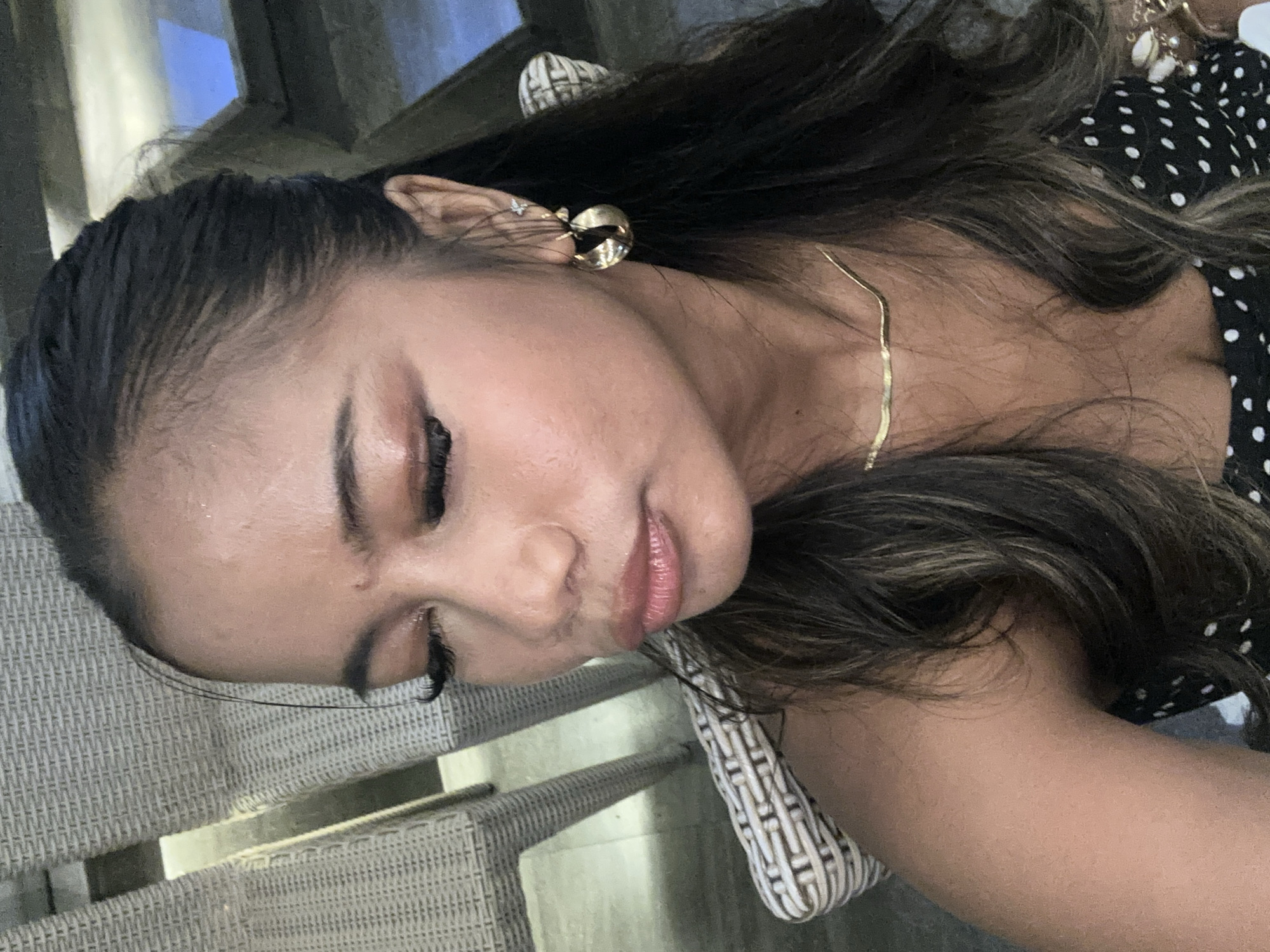 A woman with dark hair and light brown highlights, fair skin, and gold earrings is photographed indoors. She is wearing a gold necklace and a black and white polka dot top. The background includes a woven chair and a glimpse of a woven handbag. The location, Singapadu, Indonesia, cannot be determined from the image alone.