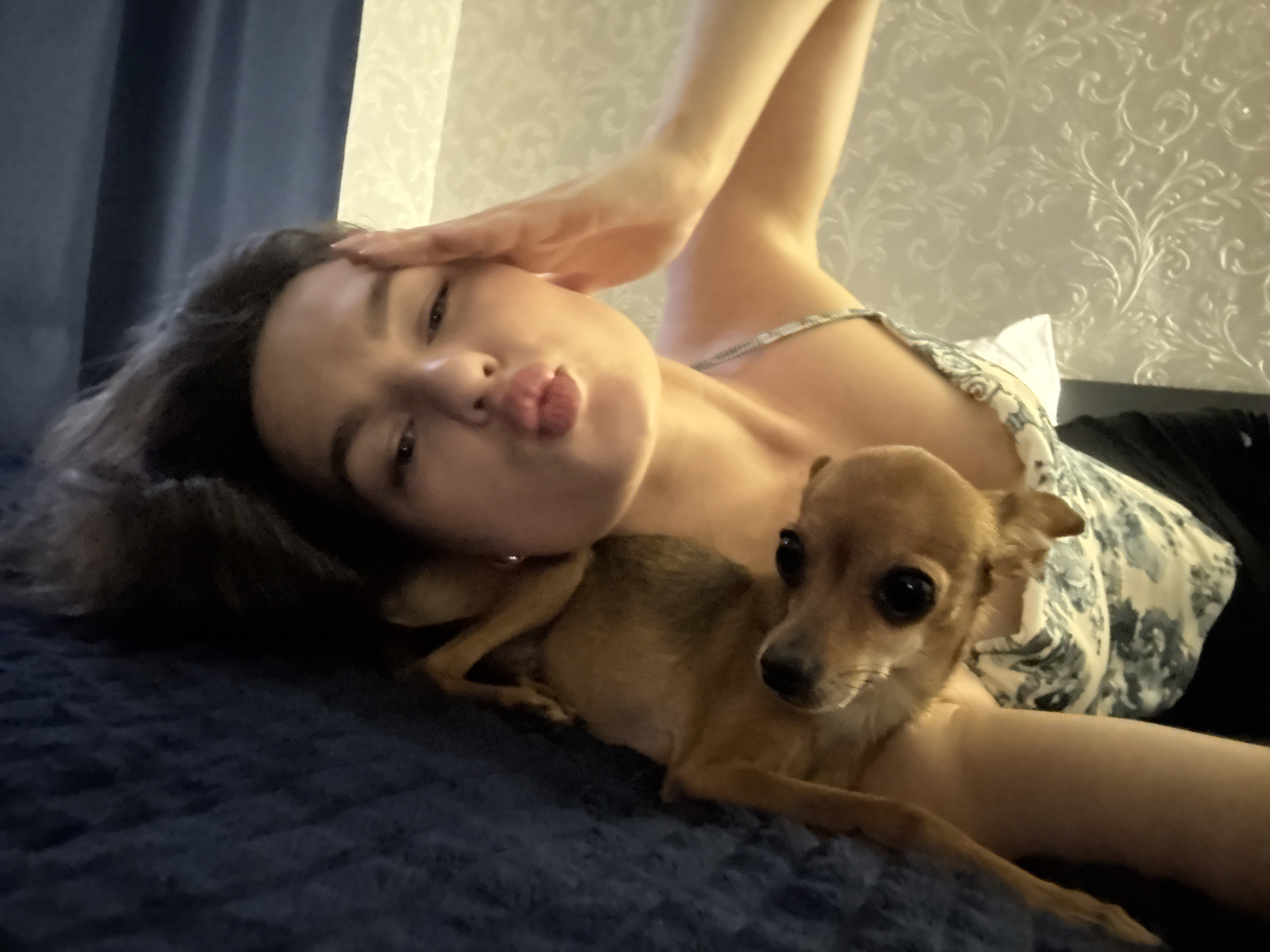 In an indoor setting, possibly a bedroom, a young woman with dark hair is lying on a dark blue textured surface. She is positioned on her left side, with her head resting on her left arm. Her right arm is raised, with her hand touching her forehead. She is looking towards the camera with her lips puckered. A small tan dog is lying on her chest, also looking towards the camera. The woman is wearing a light-colored top with a floral pattern. The background consists of a dark curtain on the left and a textured wallpaper with a floral motif on the right. The lighting suggests it is evening or indoors with artificial light. This scene does not contain any elements that would allow for determining a specific location within the city of Chaikovsky, Russia.
