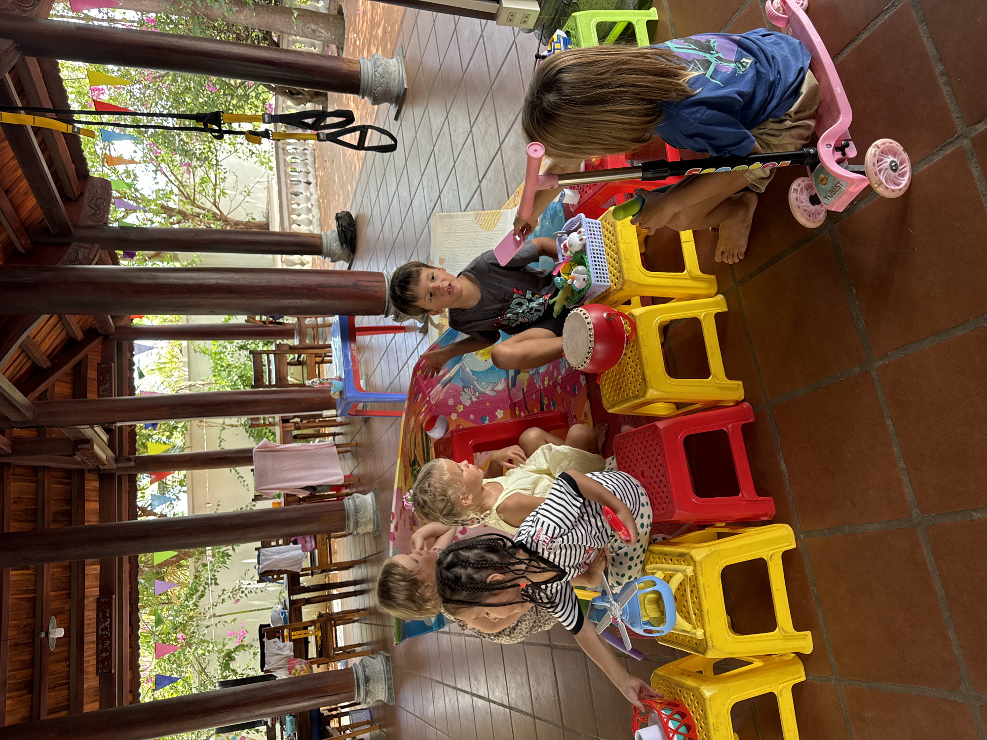 Captured at an unspecified location in Mũi Né, Vietnam, the image presents a group of children engaged in play. Five children are visible, positioned on and around a series of stacked, brightly colored plastic stools—primarily yellow and red. They interact with various toys, including a pink scooter, a drum, and an assortment of smaller items contained in a plastic basket. The setting appears to be an outdoor, covered area, characterized by a tiled floor and robust wooden columns supporting the structure. Through the columns, glimpses of foliage, colorful pennants, and domestic objects such as chairs and laundry are visible, suggesting the space is part of a residence or community center. The perspective is slightly angled, with the children and stools occupying the central foreground.