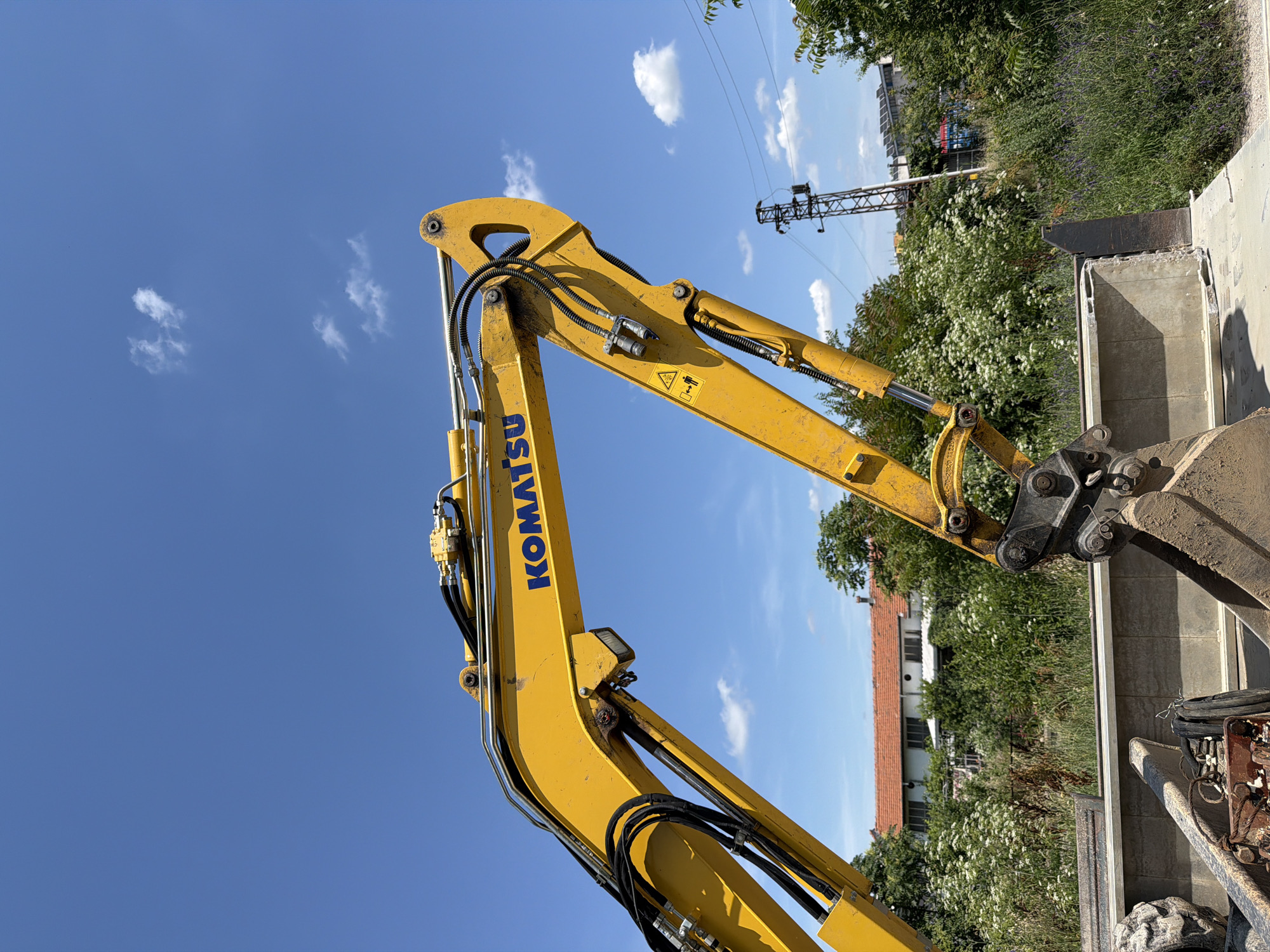 An outdoor, eye-level, medium shot captures the yellow articulated arm of a Komatsu excavator against a bright blue sky with scattered white clouds. The excavator arm, featuring hydraulic hoses and fittings, dominates the upper portion of the frame. The word "KOMATSU" is emblazoned in blue on the arm. Below the arm, a dense green foliage of trees and bushes is visible, interspersed with some white wildflowers. In the middle ground, a building with a red-tiled roof is partially obscured by the vegetation. In the background, a lattice-like industrial tower and other buildings can be seen, along with power lines. The foreground shows the lower section of the excavator, including its bucket and part of a tire. The scene appears to be a construction site or an area undergoing some form of land clearing or development.