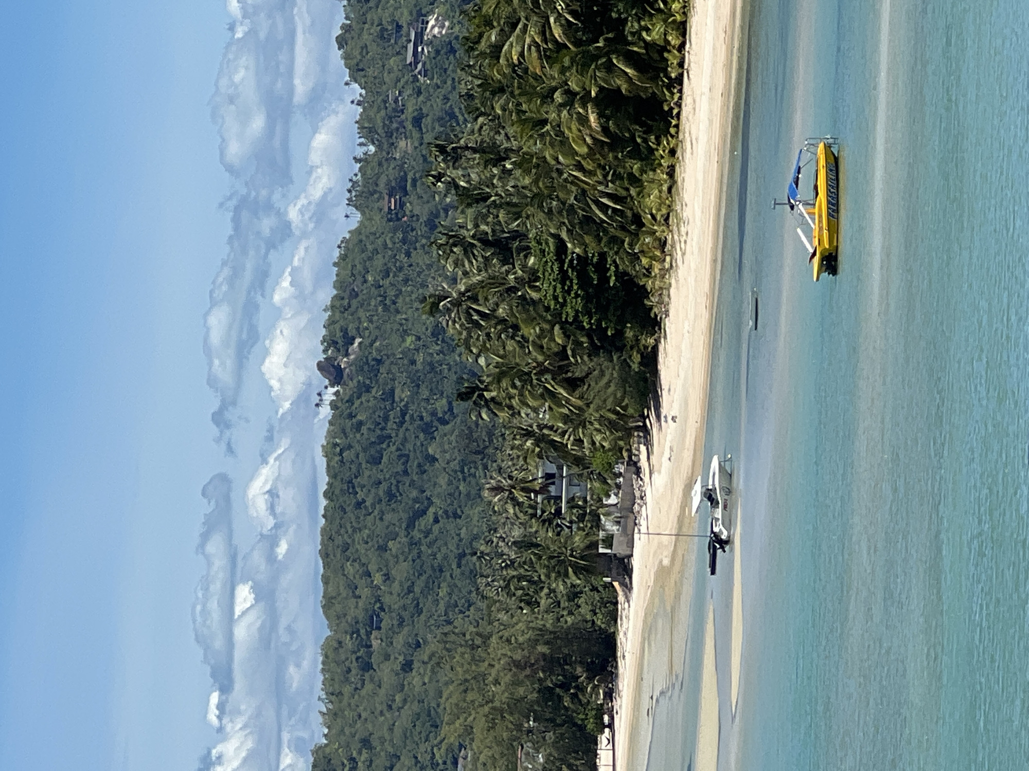 This is a high-angle, medium shot of a bay in Baan Hua Thanon, Thailand. The water is a clear turquoise, with some lighter sandbars visible beneath the surface. Two boats are in the water: a white boat with a canopy is anchored in the shallows near the beach, and a yellow boat with a blue canopy, labeled "PARASAILING," is floating in deeper water. The beach is sandy and lined with palm trees and lush green vegetation. In the background, a densely forested hill rises, with several buildings visible among the trees and on the higher slopes. The sky is bright blue with scattered white clouds. The scene depicts a tranquil tropical coastal environment.