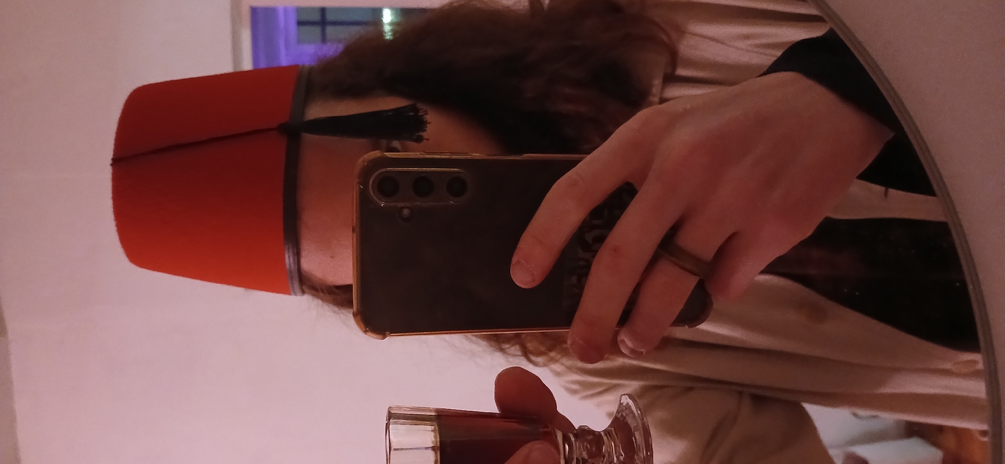 A mirrored selfie of a person wearing a red fez with a black tassel. The person is holding a smartphone in their right hand, obscuring their face in the mirror. They are wearing a brown ring on their ring finger. In their left hand, they are holding a glass containing a dark liquid. The background is out of focus, showing a glimpse of a cream-colored fabric and a window with a purple light. The location is Unknown, Unknown.