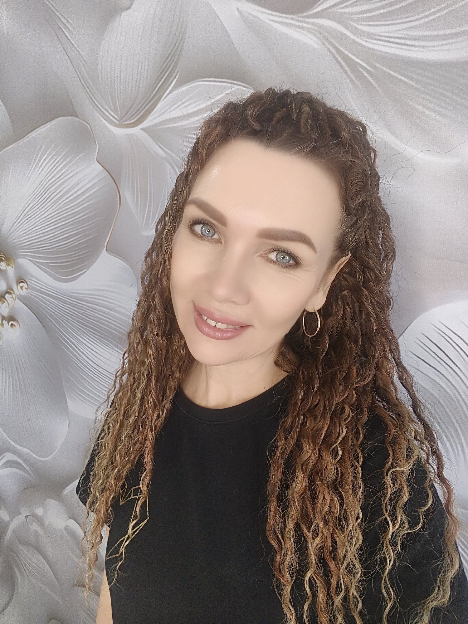 A portrait of a woman with long, curly, light brown hair wearing a black t-shirt and gold hoop earrings. She has fair skin and blue eyes and is smiling gently at the camera. The background is a white and gray floral pattern. The location appears to be indoors. The city is Unknown, Unknown.