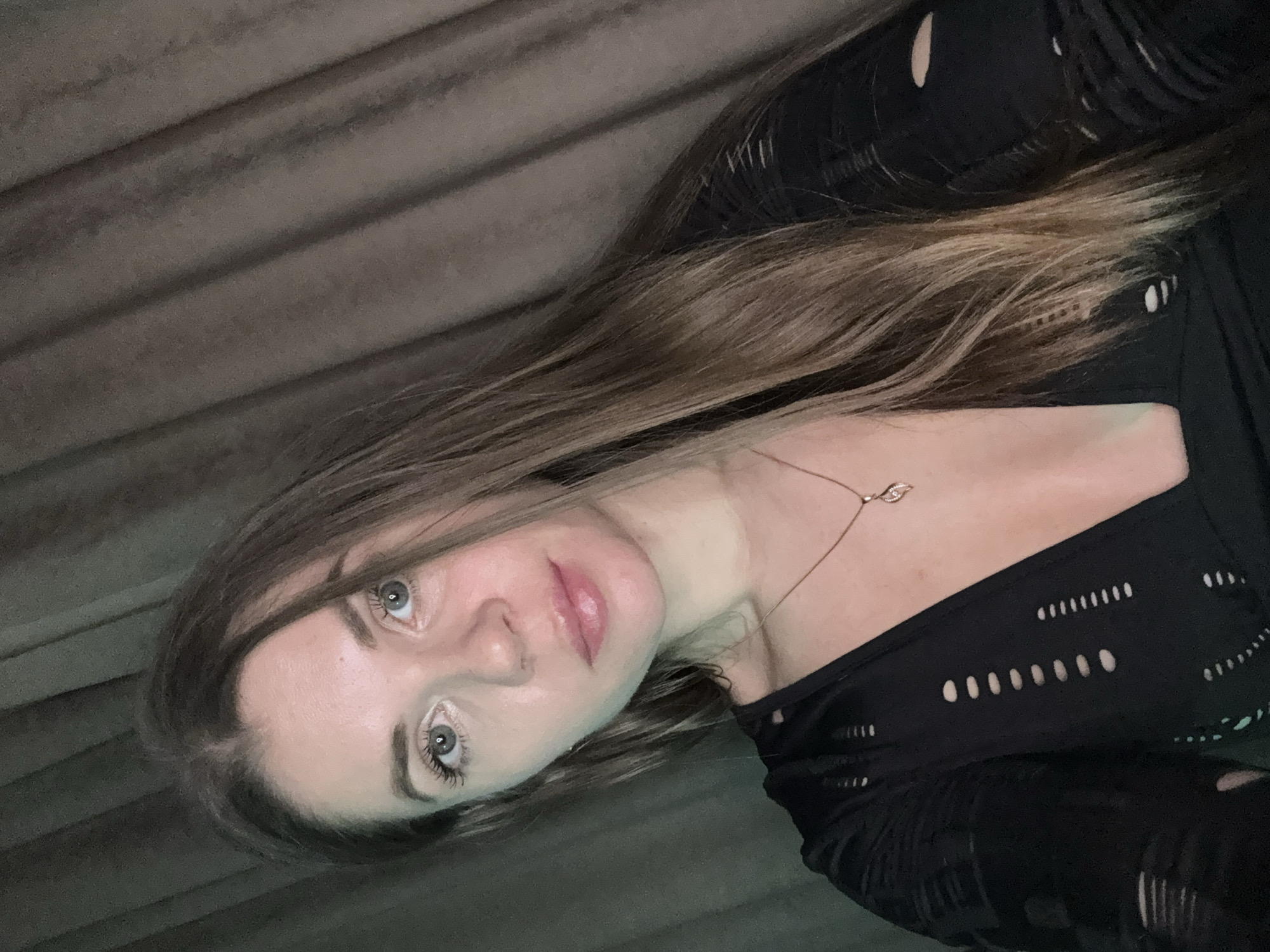 A woman is centered in the frame, her head tilted to the left. She has light skin, light eyes, and light brown hair. A necklace is visible on her neck. She wears a black top with small, oval-shaped cutouts. Behind her is a gray, textured background suggestive of a curtain or wall. No information is present to determine if the image was taken in Batumi, Georgia.