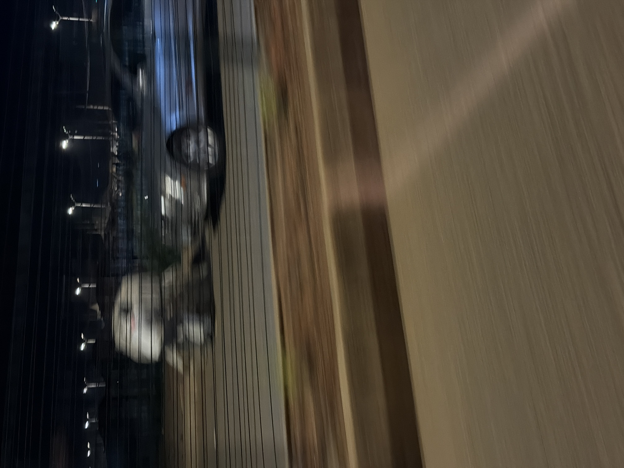 A silver sedan is in motion at night on a road, with horizontal lines obscuring the background. In the foreground, a large, light-colored, amorphous object, possibly a person in bulky clothing or a large piece of fabric, is also in motion. The streetlights in the background are blurred due to motion. The scene appears to be in Bukuru, Nigeria.