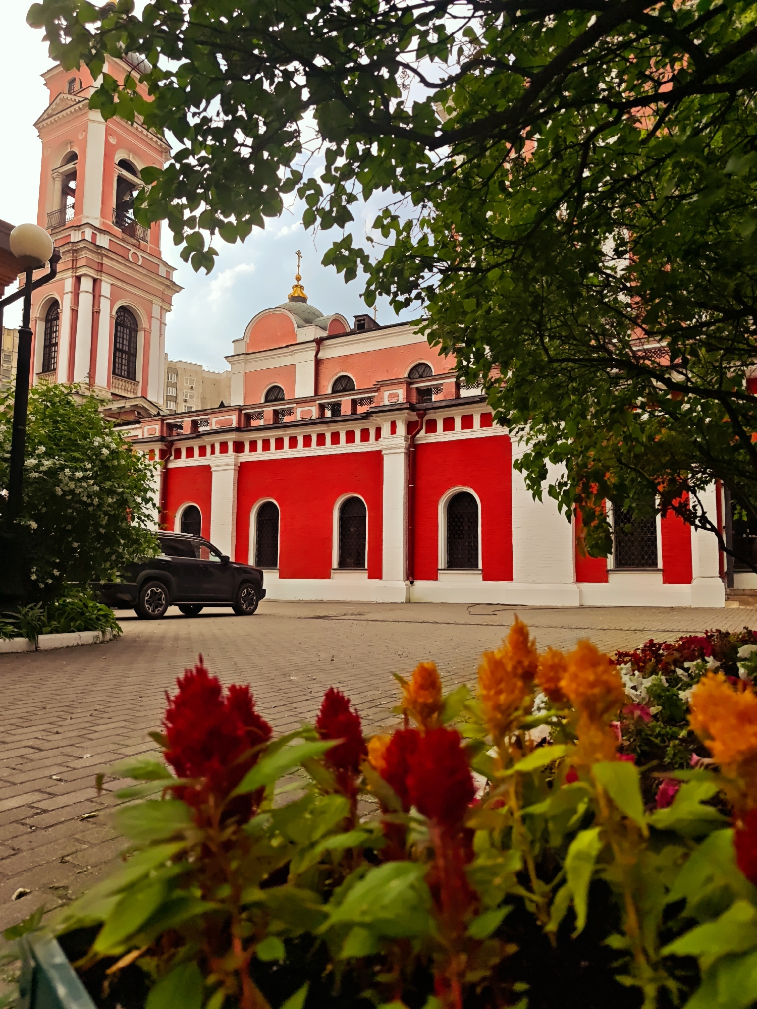 This is a photograph of the Church of St. Nicholas the Wonderworker in Khamovniki, located in Moscow, Russia. The image shows a red and white church building with a pink bell tower. The church has arched windows and a golden dome. In the foreground, there are red and orange flowers in a planter. A black SUV is parked on a brick-paved walkway. Trees with green leaves partially obscure the view of the church. The sky is partly cloudy.