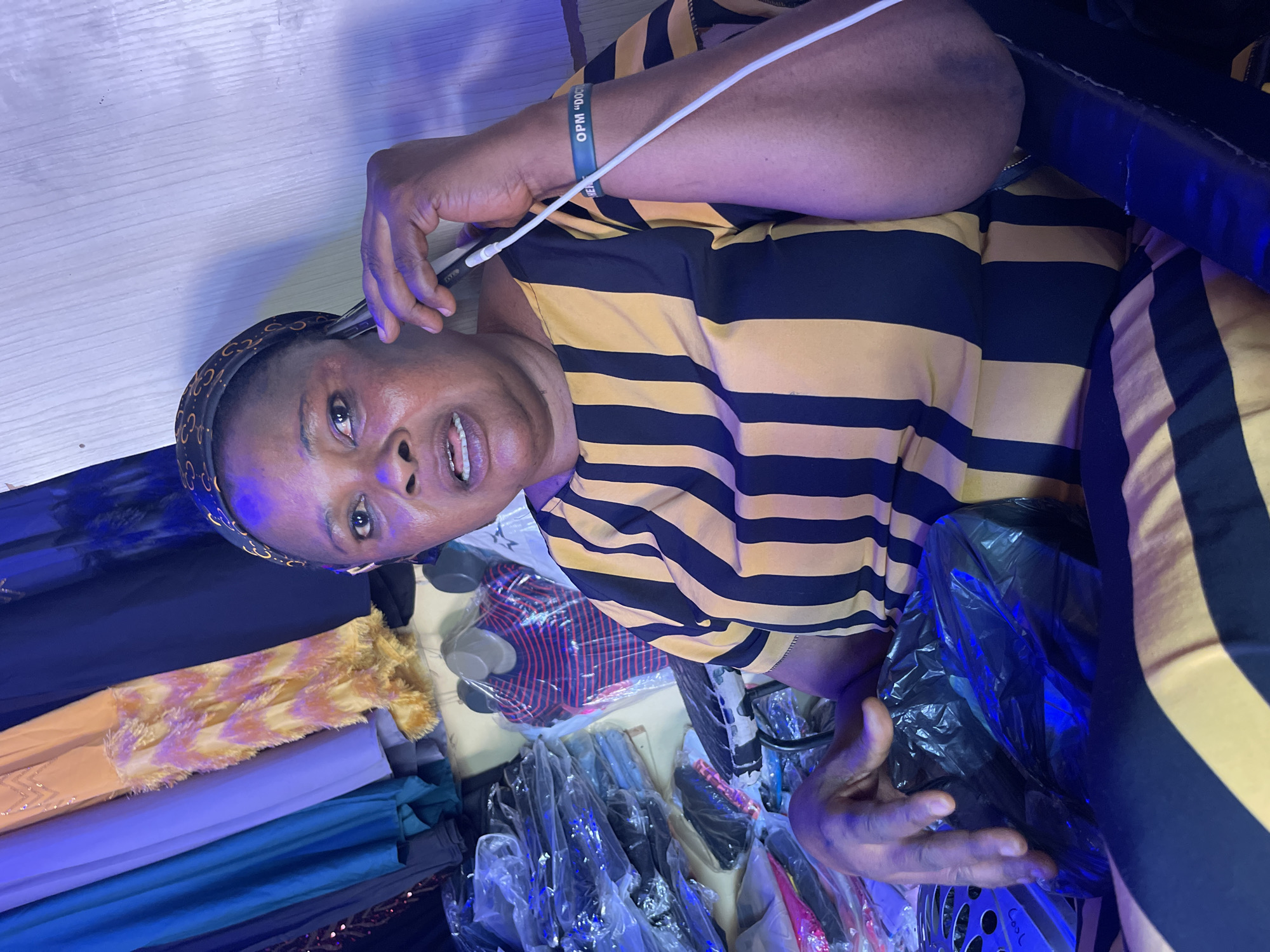 An older woman wearing a striped yellow and black shirt, a black patterned headscarf, and a green wristband is seated amidst piles of fabrics in clear plastic packaging. She is holding a white cable near her face, possibly adjusting a hearing aid. Various colored fabrics, including blues, golds, and purples, are visible. The location is assumed to be Samaru, Nigeria, based on provided information.