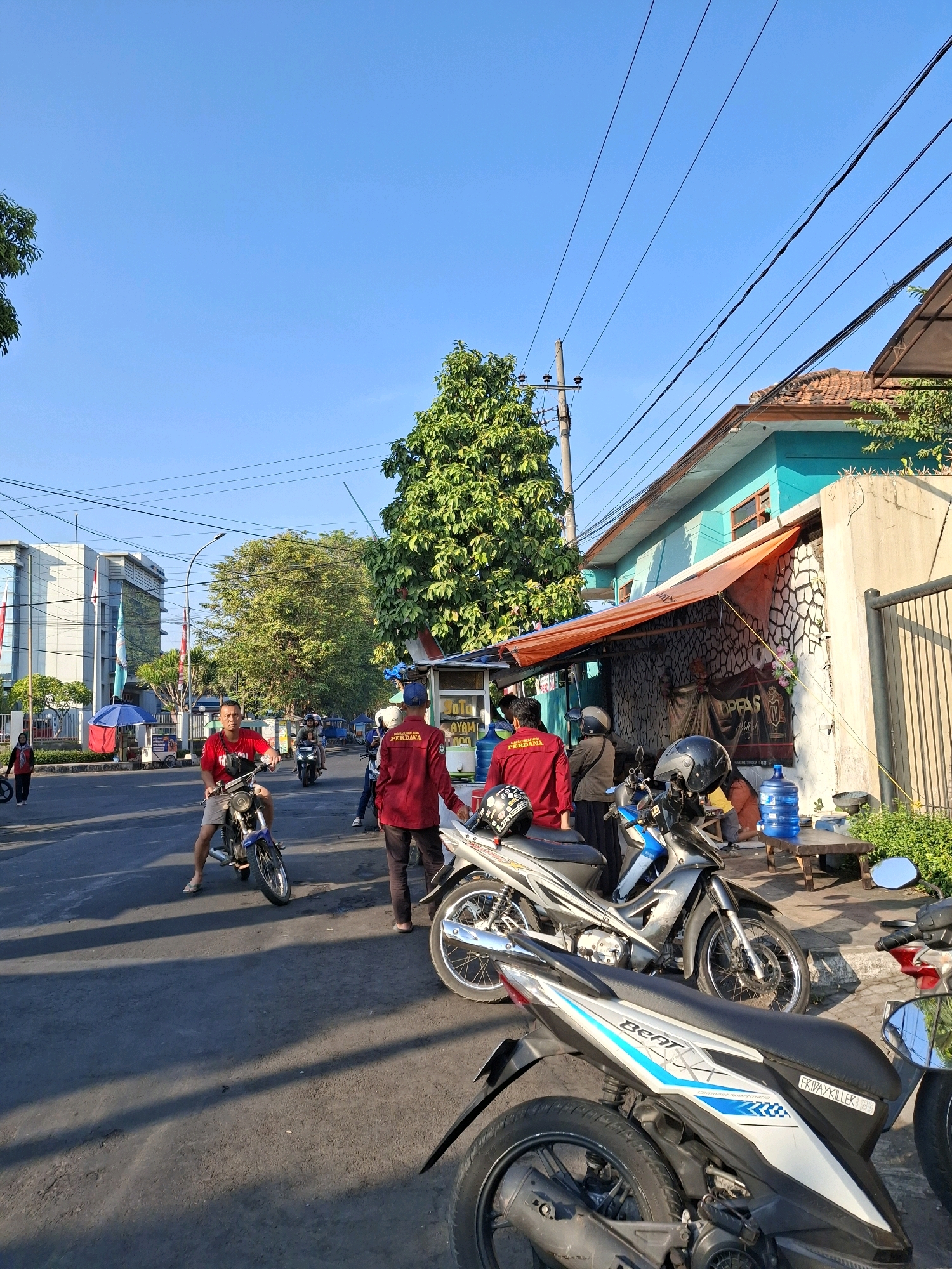 The image depicts a street scene in Lumajang, Indonesia, likely during the daytime given the bright sunlight and clear blue sky. In the foreground, several motorcycles are parked. One of these motorcycles has a sticker that reads "FRIPAYKILLER". A man in a red shirt is riding a blue and black motorcycle, and another man in a red shirt with "PERDANA" written on the back is standing near a food stall. The food stall has a blue water dispenser and a wooden bench with a metal basin on it. The stall is covered by an orange awning. Behind the stall, a turquoise-colored building with wooden window frames is visible. Further in the background, there are trees, a building with glass windows, and more motorcycles. The street is paved, and shadows are cast on it from the surrounding objects. There are electrical wires crisscrossing the sky. The overall impression is of a busy, ordinary day in a street market or commercial area.