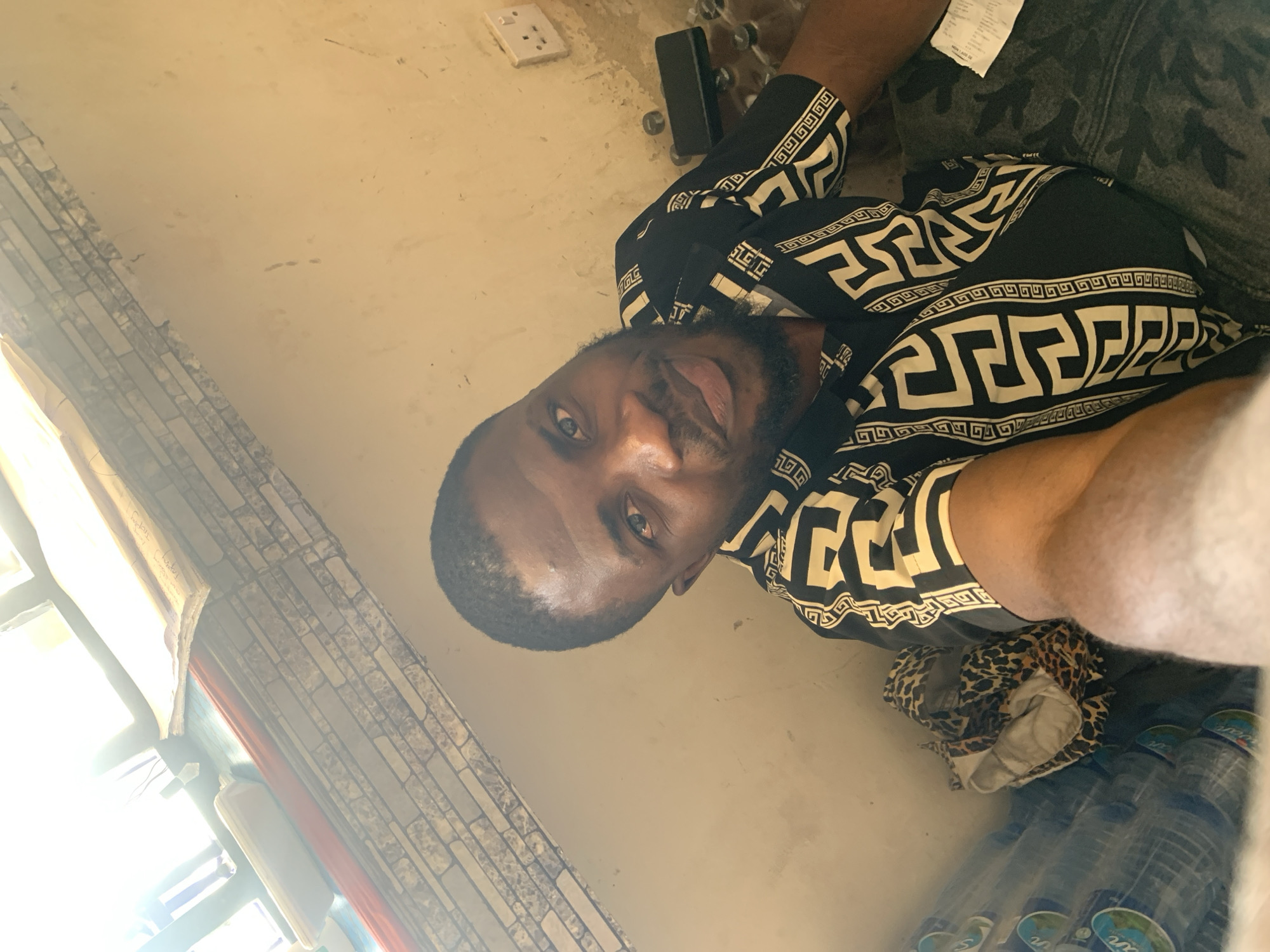 A man, appearing to be of African descent, is pictured in a close-up, selfie-style shot. He is wearing a black shirt with a geometric white pattern. His expression is relaxed, with a slight smile. The background suggests an indoor setting, possibly a room with a light-colored, textured wall. To the left, there are several plastic bottles of water, and a small, leopard-print fabric item. To the right of the man, a black rectangular object, possibly a phone, rests on a surface, and a white electrical outlet is visible on the wall. The lighting is bright, with a strong light source coming from the top left, possibly a window. The man is seated, with his right arm extended towards the camera. The setting appears to be in New Stadium, Nigeria.
