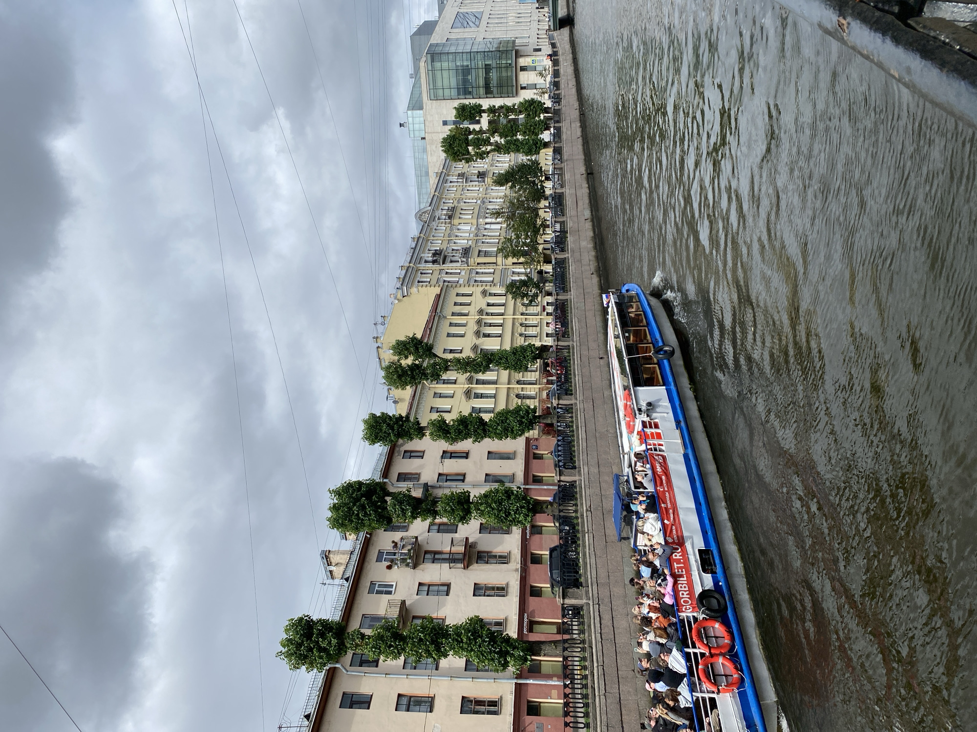 This image depicts a canal in Saint Petersburg, Russia, under a cloudy sky. A tour boat, filled with passengers, is navigating the canal, its side emblazoned with the text "GORBILET.RU". The boat is passing by a row of historic buildings and a modern glass-fronted structure, all situated along the canal's embankment. Trees line the walkway in front of the buildings, and a black metal railing separates the embankment from the water. Several cars are parked along the street behind the railing. The water in the canal is rippled, reflecting the overcast sky and surrounding architecture.