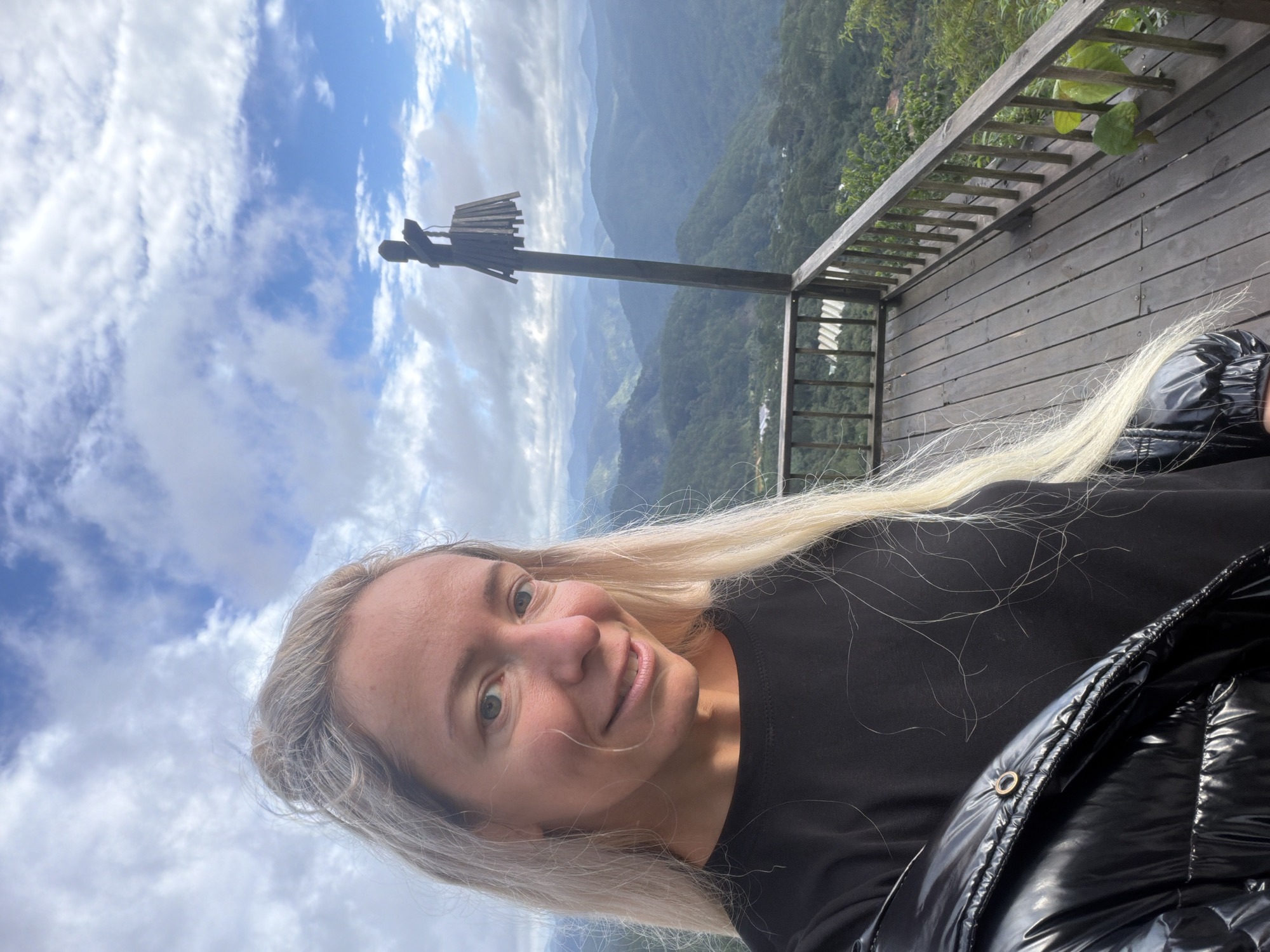 A medium shot selfie shows a fair-skinned woman with blonde hair pulled back and long strands framing her face. She is wearing a black t-shirt and a shiny black puffer jacket. She is smiling and looking at the camera. The background is a panoramic view of lush green mountains under a cloudy blue sky. The woman is standing on a wooden balcony with a railing. A dark wooden pole with a wooden structure resembling a lantern hangs from it is positioned to the right of the woman. In the distance, several layers of mountains fade into the haze, and glimpses of buildings and greenhouses can be seen nestled among the trees. The location is identified as a viewpoint in the city of Phát Chi, Vietnam.