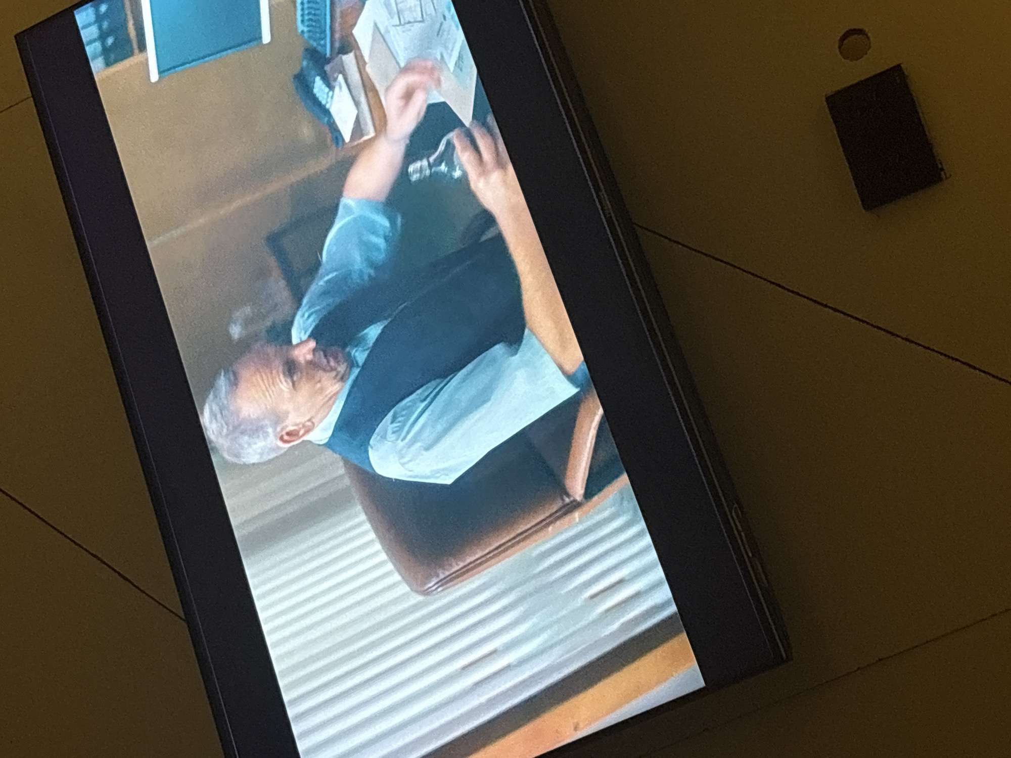 The image shows a television screen displaying a scene of an older man sitting at a desk in an office. He is wearing a light blue shirt and a dark vest. He appears to be looking down at something on the desk, with his hands raised. The desk has some papers, a calculator, and other office items on it. The office has a wooden desk, a brown leather chair, and striped wall paneling. Adjacent to the television screen on the right side of the image, on what appears to be a tan-colored surface, there is a small round object and a rectangular dark object. The photograph was not taken in Iddo Tudunwada, Nigeria. There is no evidence to support this.
