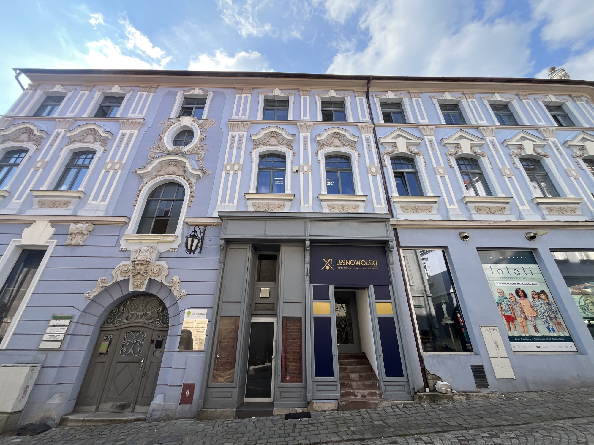 The image shows a wide-angle, eye-level outdoor shot of a light purple, ornate building facade on a cobblestone street. The building has multiple stories with numerous windows. The architectural style features decorative white and tan embellishments around the windows and along the facade, including pilasters and scrolled motifs.
On the ground floor, there are several entrances and storefronts. To the left, there is a large, arched wooden door with ornate metalwork. Above this door, there are signs in Polish, one with a green border. To the right of this door, there is a gray doorway with visible brickwork behind glass panels, and a smaller entrance with a white door. Further to the right, there is a prominent storefront with a blue awning displaying the text "LEŚNOWOLSKI" in white and a gold sewing needle logo. This entrance has a set of stairs leading up to a dark blue door. Adjacent to this storefront, to the far right, is a display window with a large poster featuring children modeling clothing, with the text "lalaLi" and other Polish words. The poster has a red border at the top and a white border at the bottom.
The sky above is blue with scattered white clouds. The building's facade appears to be leaning slightly to the right, causing a distortion in the perspective. The cobblestone street in the foreground is well-maintained. There are no people visible in the image.