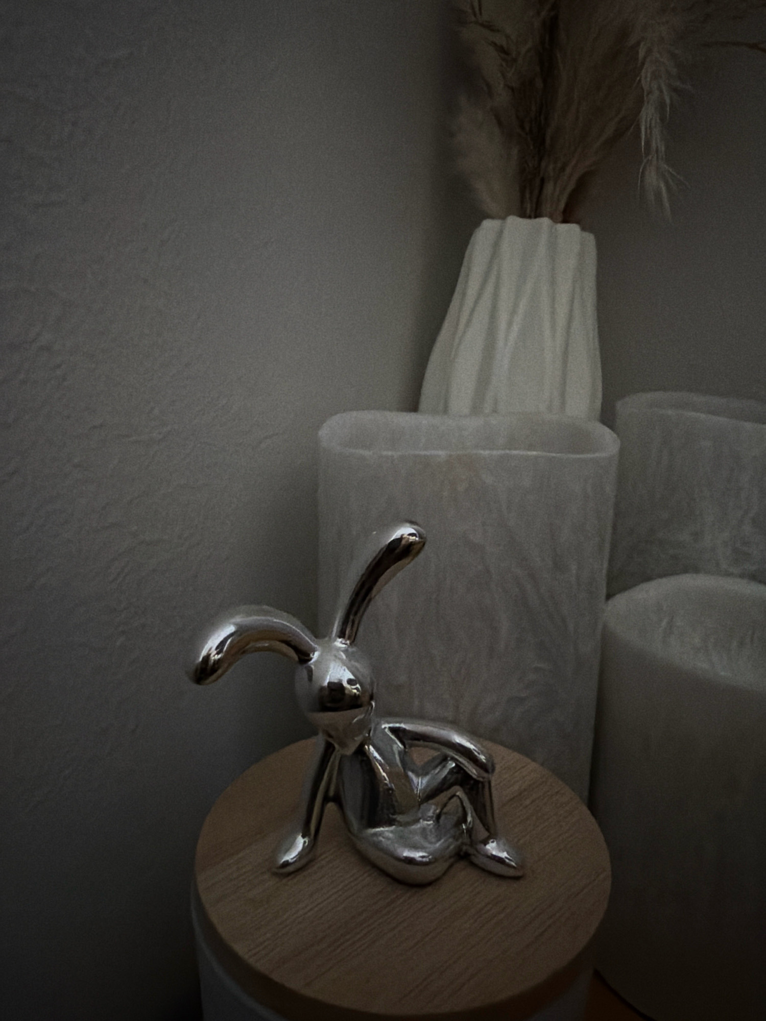 A silver rabbit figurine sits on a wooden surface, which rests on a white cylindrical object. In the background, there are three large, translucent white cylindrical objects, and a white vase holding dried grass stalks. The scene is dimly lit, with a textured grey wall in the background. The image does not provide enough information to determine its location in Helsinki, Finland.