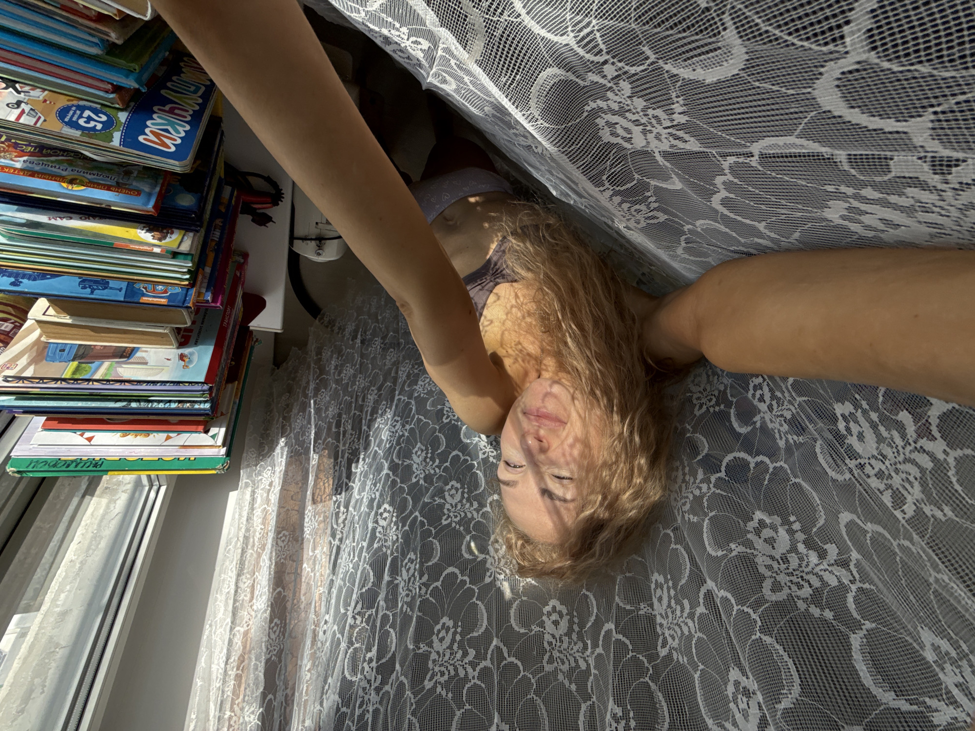 A woman with blonde hair is lying under a white lace curtain near a window and a stack of books. She appears to be looking up at the camera. The photograph was taken indoors, presumably in a private residence. The city of Chaikovsky, Russia is not discernible from the visual content of the image.