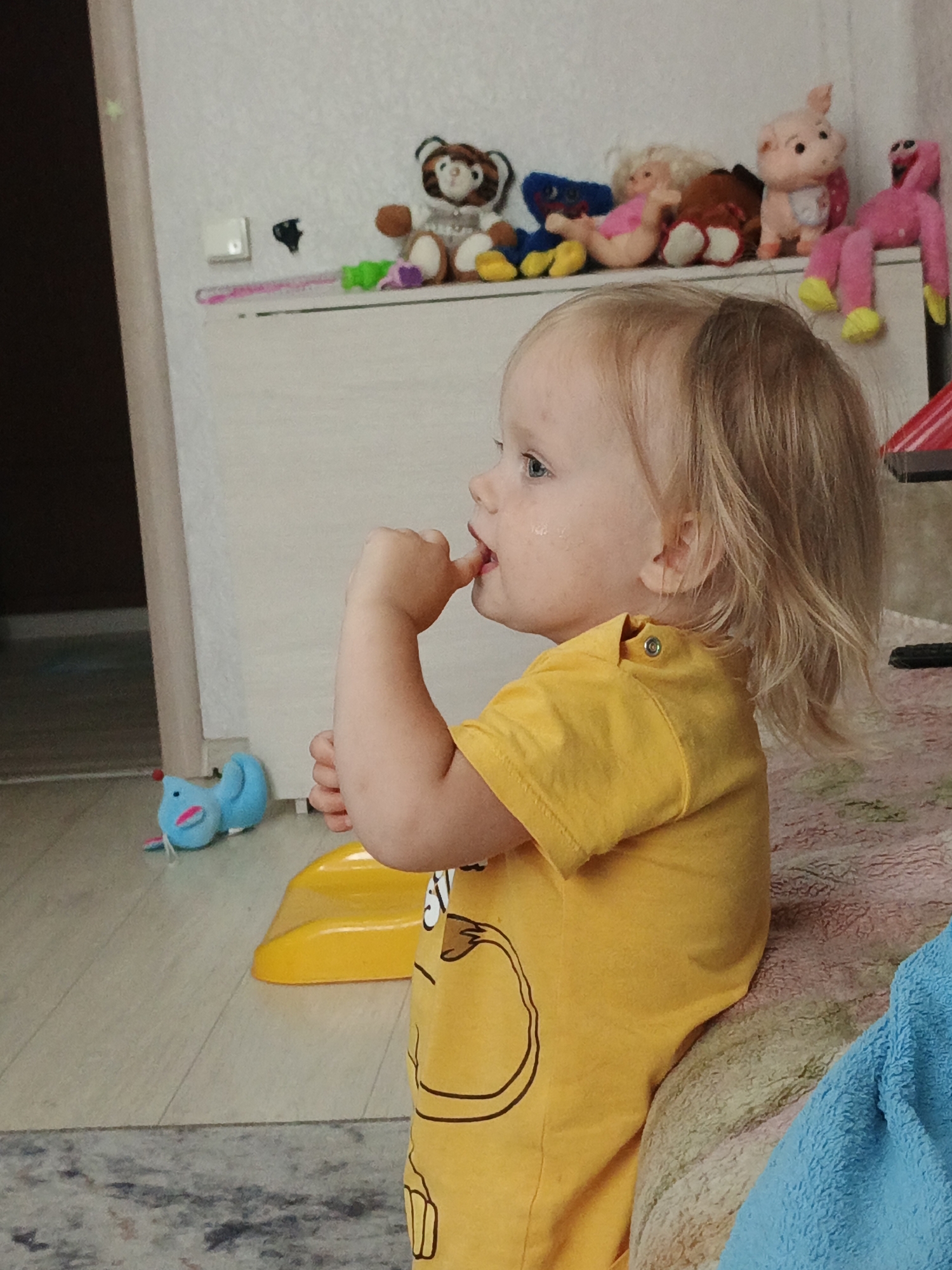 A toddler with blonde hair and blue eyes, wearing a yellow t-shirt with a black cartoon design, is sitting indoors and putting their finger in their mouth. The toddler is facing left, with their head turned towards the right. In the background, a shelf is visible with several stuffed animals, including a tiger, a pig, and a pink creature. A blue toy is on the floor to the left of the toddler, and a yellow plastic toy is in front of them. The overall environment appears to be a child's room or living space. The image does not provide any information about the city of Zlatoust, Russia.