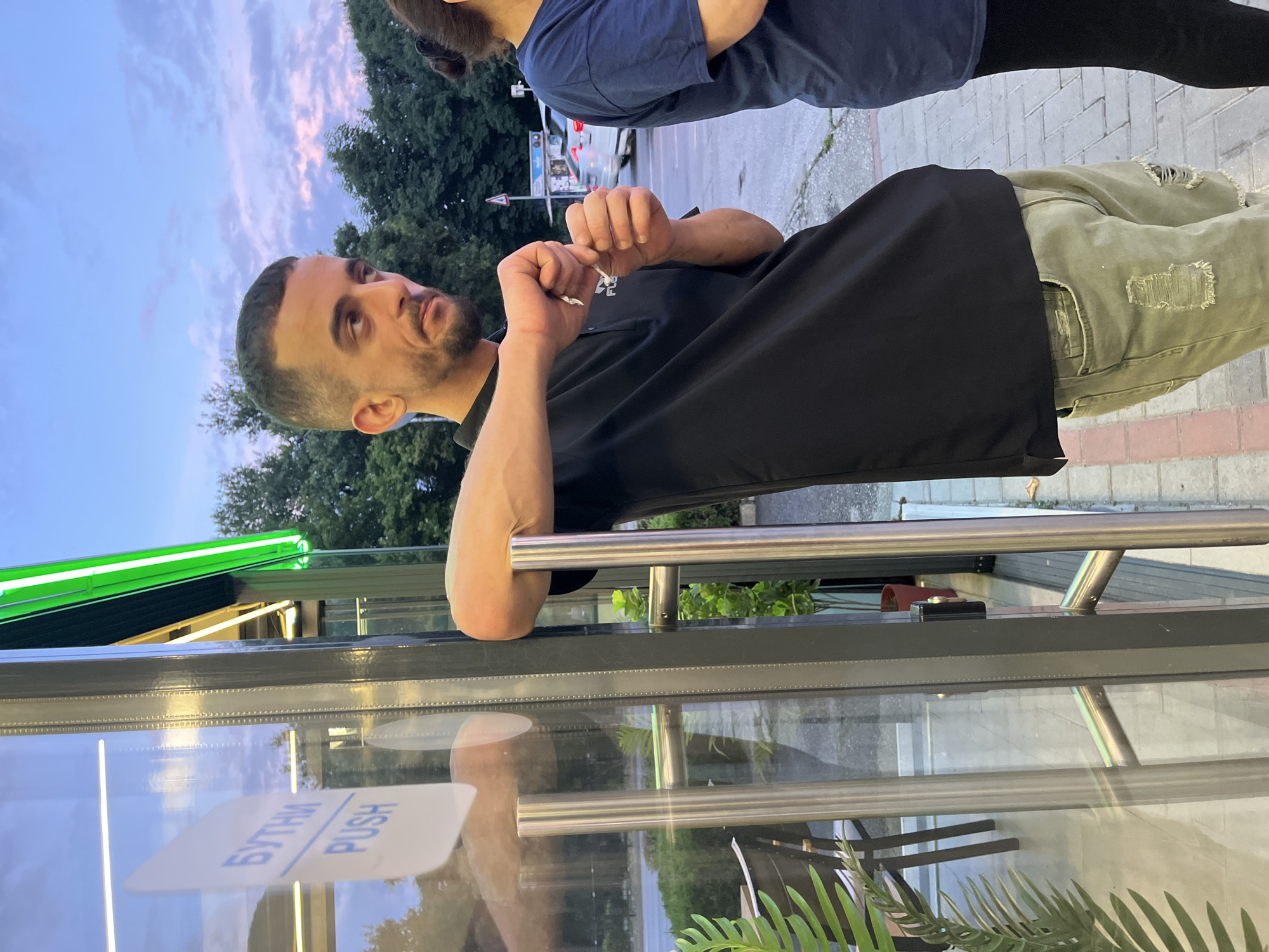 In Plovdiv, Bulgaria, a man with short dark hair and a light beard is leaning on a glass door with silver handles. He is wearing a black polo shirt and ripped olive green jeans. He is looking to his right and holding a small white object in his hands, which he appears to be tearing apart. Next to him, a woman with dark hair tied up is standing, wearing a dark blue t-shirt. The door has a sticker with Cyrillic text and the word "PUSH" in English. In the background, trees and a street with cars are visible under a twilight sky.