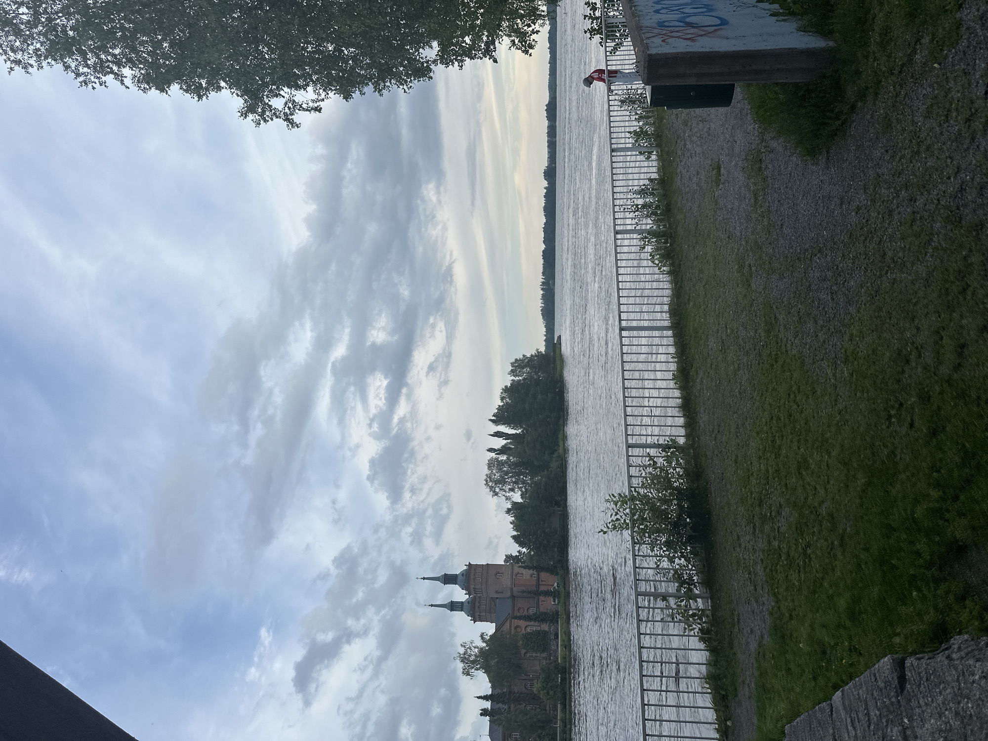 The image depicts a waterfront scene in Vammala, Finland, featuring a large brick building with two prominent towers on the far bank of a wide river. The sky is cloudy with varying shades of grey and white. In the foreground, a metal railing separates a grassy area from the river. A person wearing a red and white shirt is standing by the railing on the right side of the image, facing away from the camera and towards the water. To the right of the person, a trash bin is visible, and a concrete structure with graffiti is present. Trees and vegetation are scattered throughout the scene, with a dense cluster of trees behind the brick building on the opposite bank. The water appears calm with ripples.