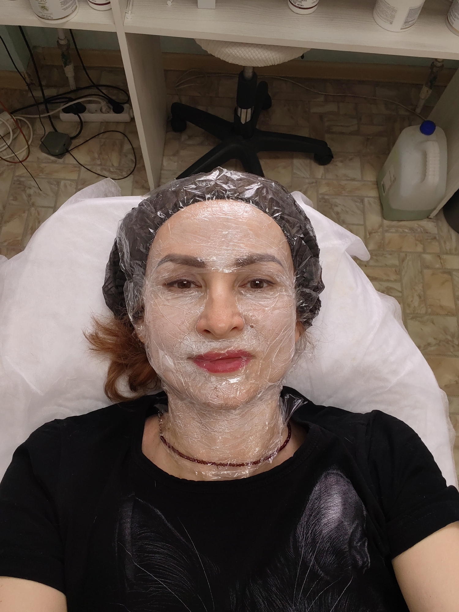 A woman lies supine on a white examination table, face covered in a translucent wrap with a dark hair cap. She wears a black t-shirt with a white design, and a beaded necklace. The environment appears to be a medical or cosmetic treatment room in Chaikovsky, Russia, evident from the style and fixtures. An office chair is visible at the head of the table. Bottles and electrical cords are on a shelf and floor. The floor tiles are patterned. A large plastic container with a blue cap is visible in the right background.