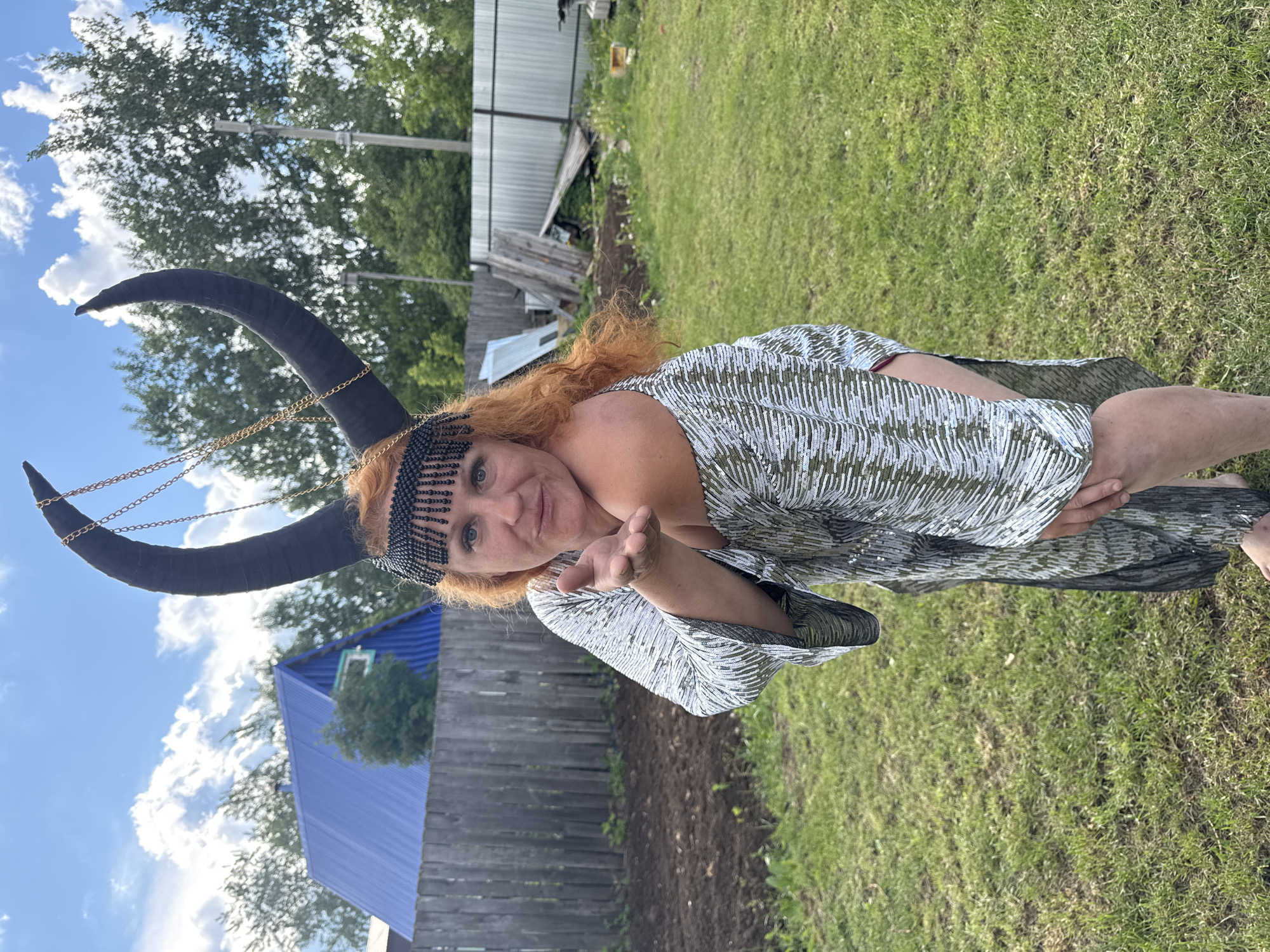 A woman with long, curly red hair is captured outdoors in a daytime setting, posing for the camera. She is wearing an elaborate headpiece featuring large, curved black horns draped with gold chains, and a black beaded fringe across her forehead. Her attire consists of a shimmering, long-sleeved dress or robe, patterned with numerous short, light-colored lines on a darker base. She extends her left hand towards the viewer, palm slightly open, with a gentle smile on her face, conveying a friendly or playful expression as she looks directly at the camera.
The background reveals a grassy area where the woman stands. Behind her is a weathered wooden fence, and further back, a building with a distinctive blue corrugated roof is visible. Various trees and greenery are scattered throughout the scene, and the ground near the fence appears to be disturbed soil, possibly a garden bed. The sky above is blue with scattered white clouds, indicating clear, sunny weather. The overall setting suggests a rural or backyard environment, located in Chaikovsky, Russia. There is no visible text in the image.