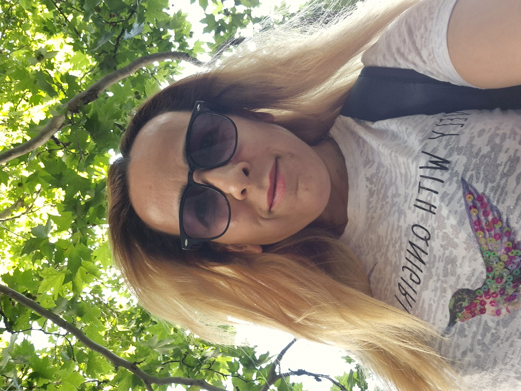 A low-angle, close-up, selfie-style photograph shows a light-skinned person with blonde hair, wearing sunglasses and a white t-shirt with a hummingbird graphic. The person is outdoors, with lush green foliage and branches from a tree visible in the background, suggesting a park or natural setting within an urban environment. The overall lighting is bright, with sunlight filtering through the leaves.