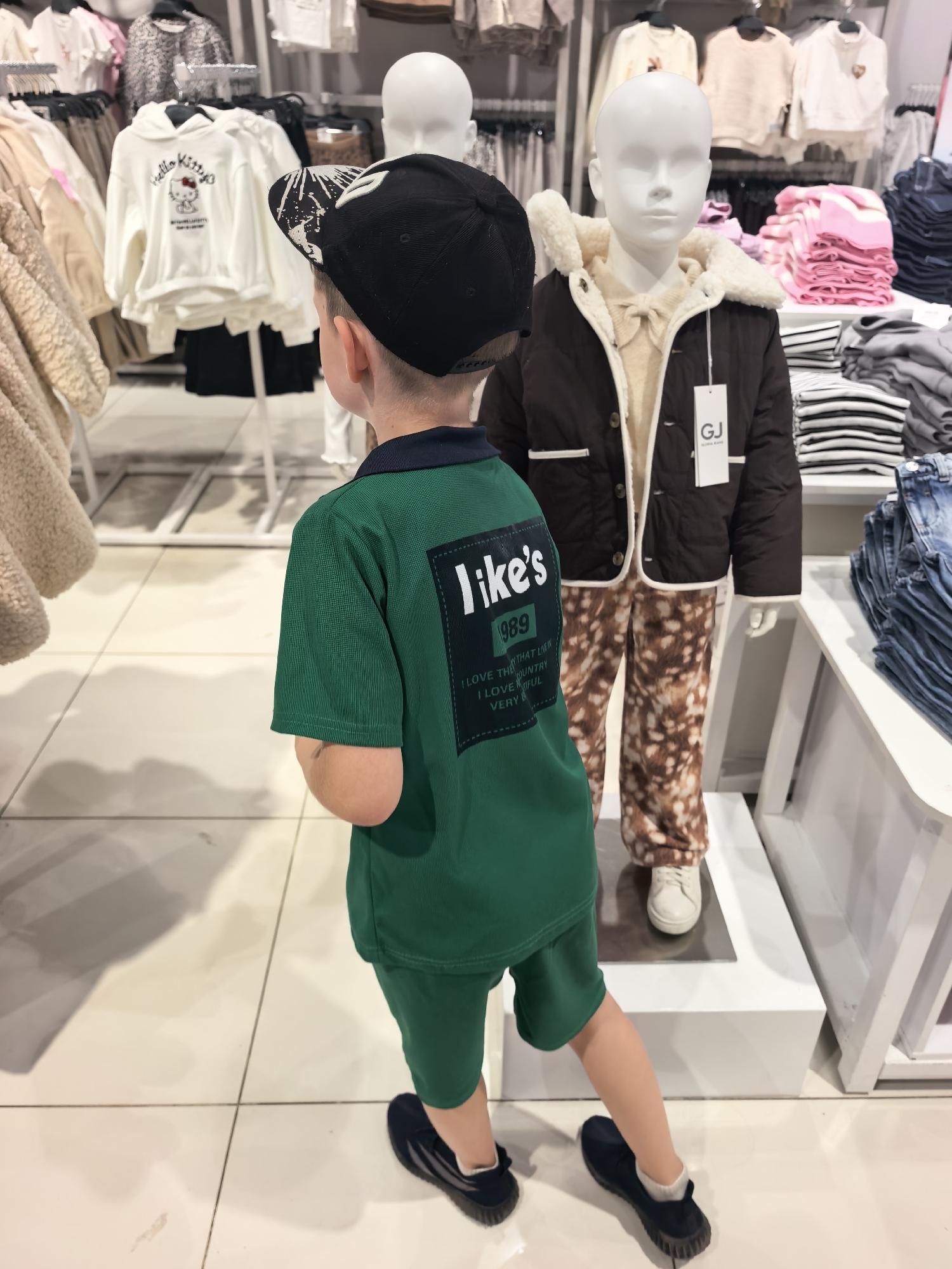 A young boy wearing a green polo shirt and shorts, a black baseball cap, and dark sneakers stands in a clothing store. The boy is facing away from the camera, looking towards a mannequin. The mannequin is dressed in a brown jacket with a fur-lined collar, a cream-colored shirt with a bow, and patterned trousers. Behind the boy and the mannequin, racks of clothes and folded stacks of clothing are visible, including white hoodies with Hello Kitty graphics, pink and white striped tops, and denim jeans. The floor is tiled with a light-colored material. The store appears to be well-lit. The image was taken in a clothing store in Sterlitamak, Russia.