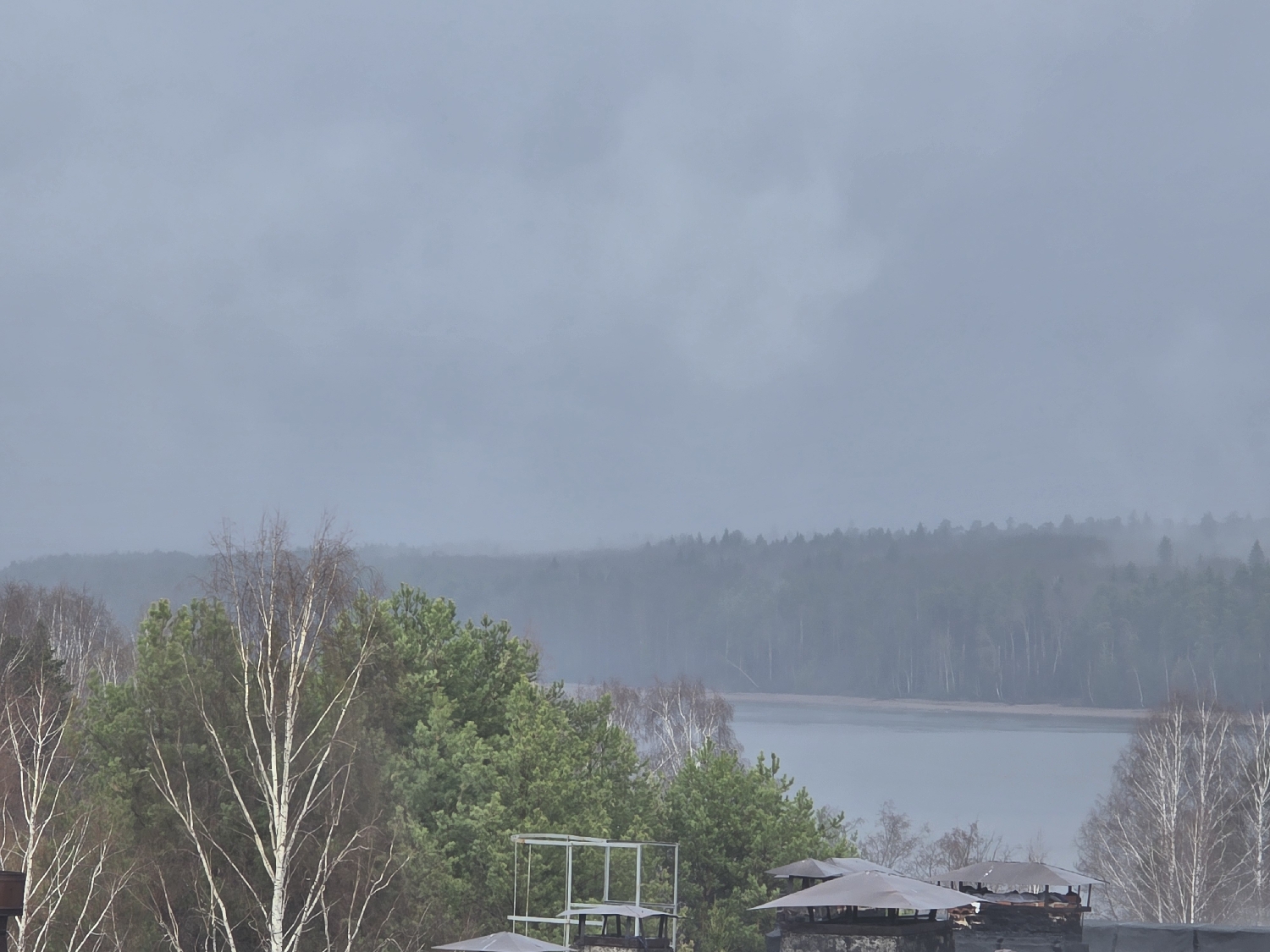 The image presents a serene, misty landscape, likely captured in Chaikovsky, Russia. The scene is dominated by a wide body of water, possibly a lake or a broad river, which stretches across the middle ground and recedes into the hazy distance. This water body is flanked by extensive, dense forests.
In the foreground, a mix of tree types is visible. Coniferous trees, identifiable by their green needles, stand alongside numerous deciduous birches. Many birch trees appear bare, indicating late autumn, winter, or early spring, though some show hints of early foliage. A prominent white-barked birch tree stands near the center-left. At the very bottom of the frame, several dark, pointed roof structures, possibly chimneys or ventilation shafts, are visible atop what appears to be a building, along with a metallic framework or scaffolding.
The sky is heavily overcast, contributing to a muted, flat lighting condition across the entire scene. A thick layer of haze or mist blankets the distant forest and the water, obscuring the horizon and lending an atmospheric, somewhat melancholic feel to the landscape. The weather appears damp and cool. No people are visible, and there is no discernible activity taking place, making it a tranquil, static view of nature. No text is present in the image.